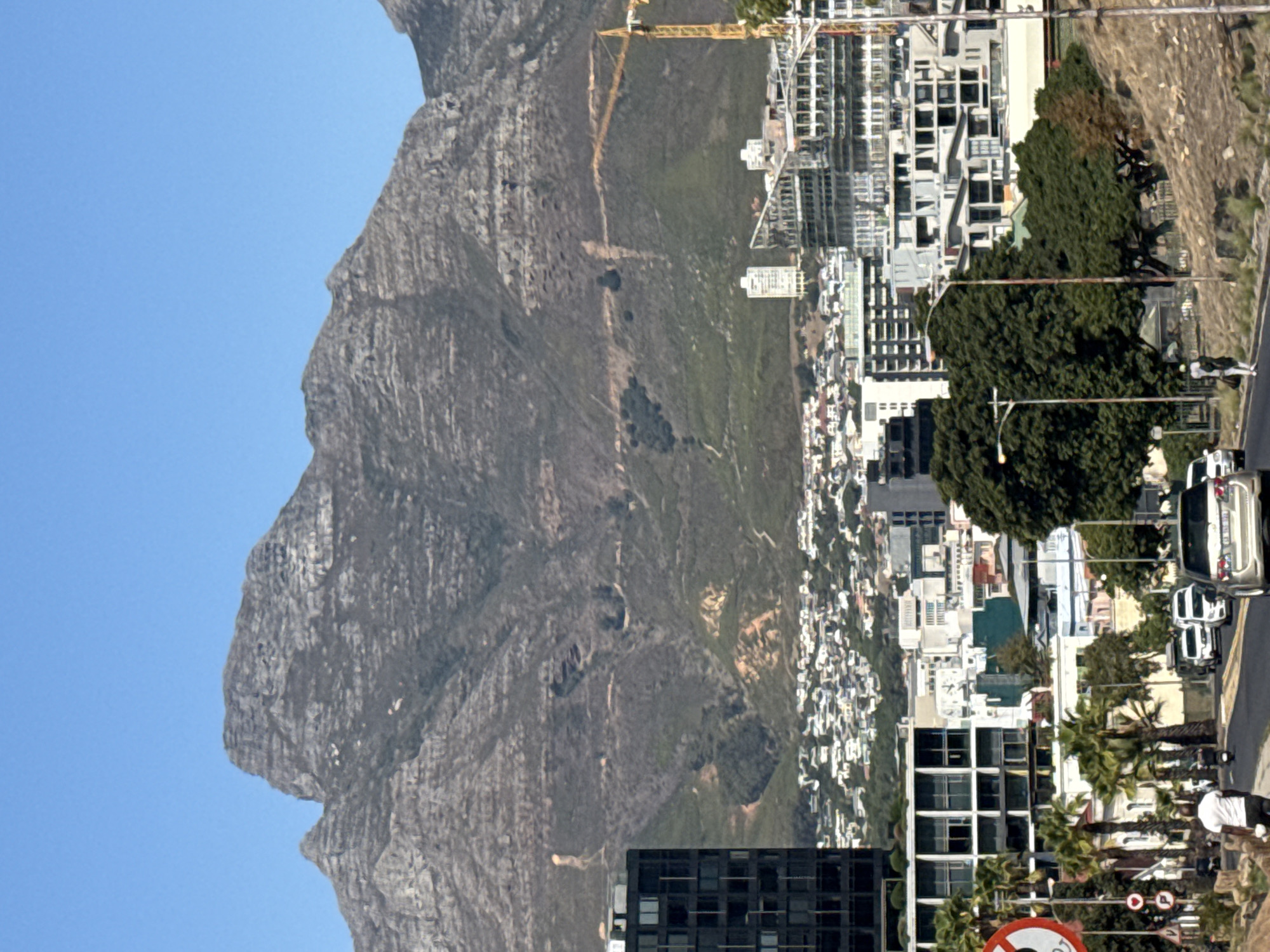 The image shows a view of Cape Town, South Africa. Dominating the left is the prominent Table Mountain, with a clear blue sky behind. Transitioning rightward, the scene depicts the mountainside, gradually giving way to dense urban development. This includes buildings of varying heights and architectural styles. A construction crane is visible. A road runs through the lower right portion of the image, with cars and pedestrians. Palm trees and other vegetation add greenery to the urban environment.
