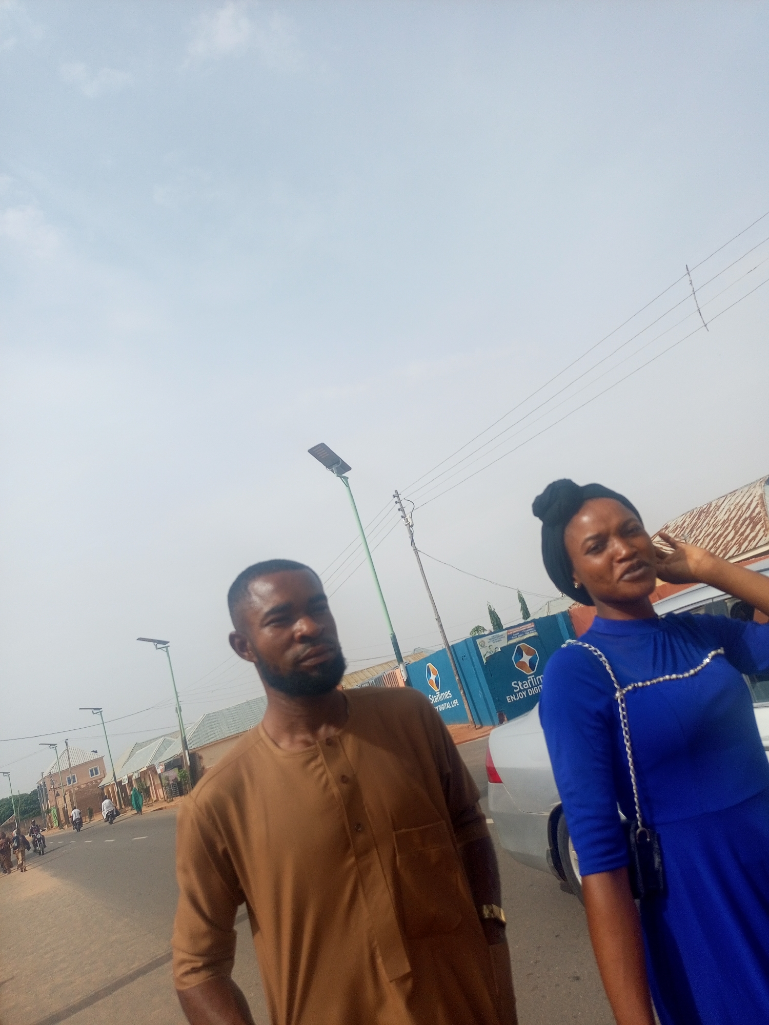 Two people stand on a paved road. A man wears a brown, long-sleeved shirt. A woman wears a blue dress with a silver chain detail. A silver car is parked near them. The background includes buildings, streetlights, and a blue wall with the "StarTimes" logo. Other people and vehicles are visible further down the road. The location is likely a street in Nigeria.