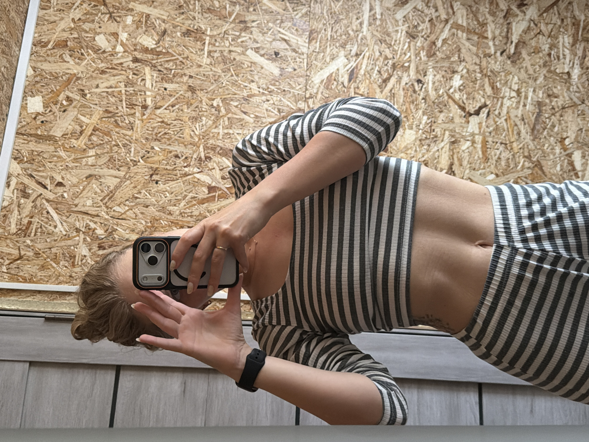 A woman is reflected in a mirror, taking a photo of herself with a smartphone that has a smiley face on the back. She is wearing a striped, form-fitting top with long sleeves and appears to be wearing striped shorts or pants as well. A black smartwatch is visible on her left wrist. The background is a wall covered in particle board. The location, Chaikovsky, Russia, is not discernible from the image.