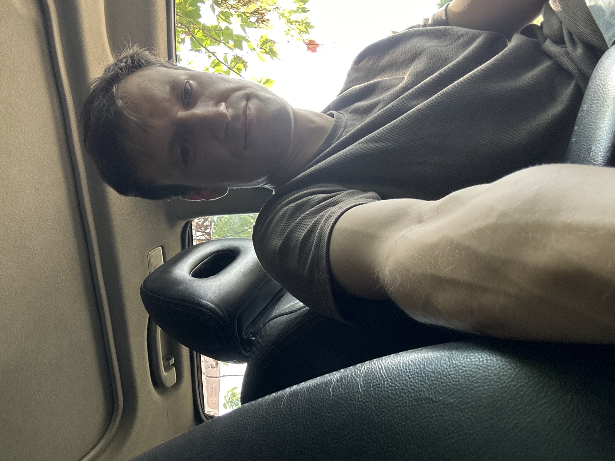 A man is sitting in the driver's seat of a vehicle, facing the camera. He is wearing a dark grey t-shirt and his right arm is extended towards the camera. The interior of the car is visible, including the headrest of the passenger seat and a portion of the sunroof. Sunlight is streaming in from the right side of the frame, illuminating the man's face and arm, and casting shadows inside the car. The background outside the car appears to be foliage and some structures. The exact location in Makiv, Ukraine cannot be determined from the image alone.