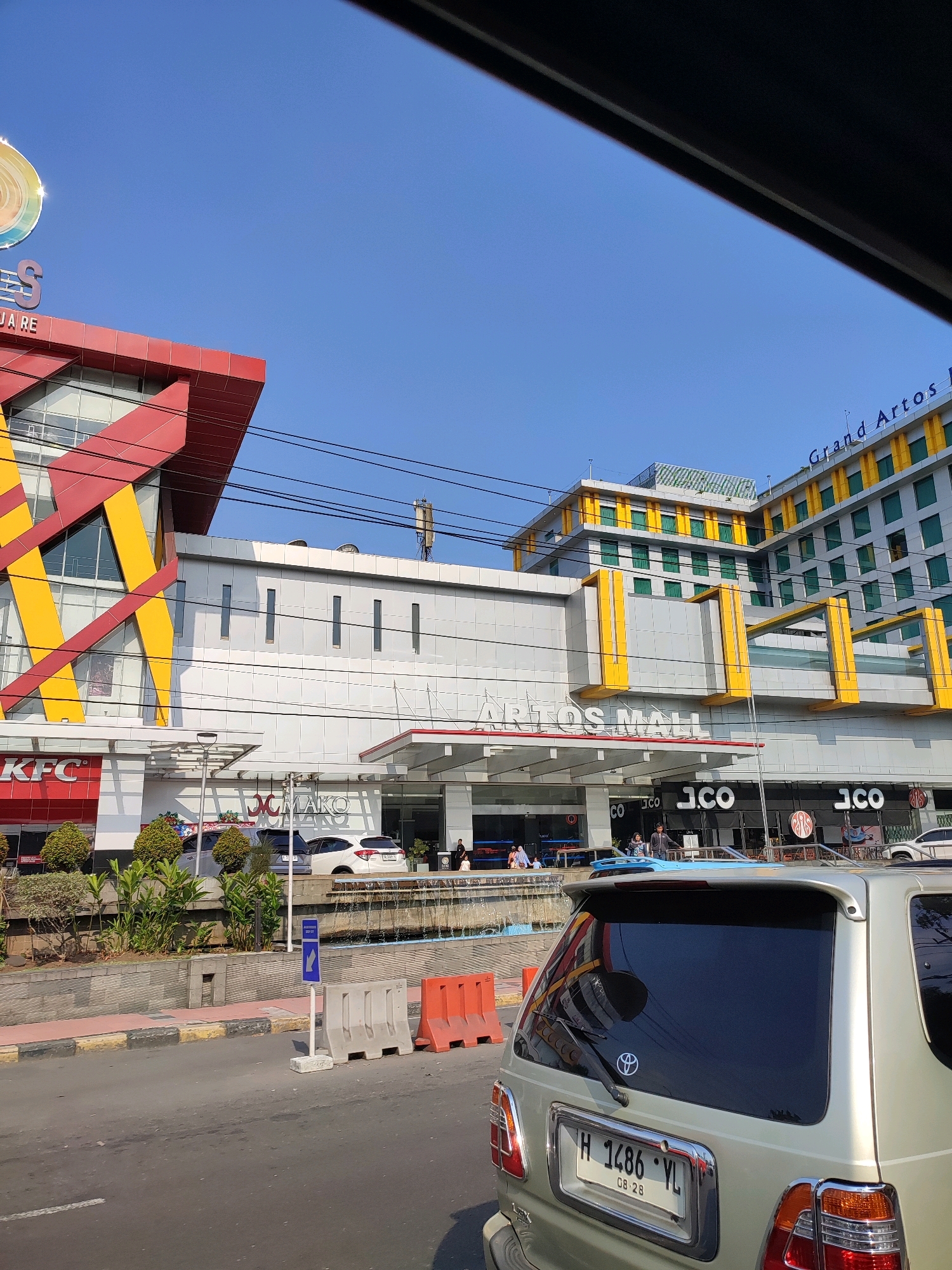 The image shows a street view of the Artos Mall and Grand Artos Hotel in Banyurojo, Indonesia. In the foreground, a silver minivan with a license plate reading "H 1486 YL" is parked. The minivan has a spoiler and tinted windows. To the left of the minivan is a section of the road with a sidewalk, orange traffic barriers, and a blue directional sign. Behind the barriers, there is a fountain and landscaping in front of the mall.
The Artos Mall has a red and yellow architectural design on one section and a white facade with vertical windows on another. The text "ARTOS MALL" is visible above the entrance. Several shops are visible at the ground level, including "KFC" and "J.CO".
The Grand Artos Hotel is a tall building with a green facade and yellow accents. The text "Grand Artos H" is visible on the hotel building. The sky is clear blue. A few people are visible walking in front of the mall entrance and near the shops. The overall scene depicts a commercial and hotel complex on a sunny day.