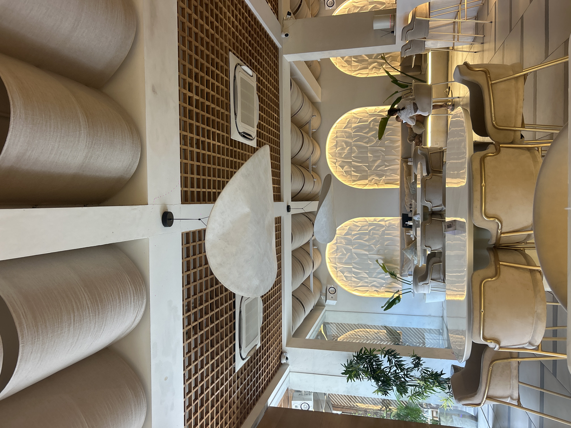 The image shows the interior of a restaurant in North Kuta, Indonesia. The ceiling features a unique design with curved fabric panels and a wooden lattice structure. A large, modern fan hangs from the ceiling. In the foreground, a white, circular table is surrounded by plush, beige chairs with gold-colored legs. Further back, there are more tables and chairs, as well as a bar area with high stools. The walls are decorated with textured panels that are illuminated by warm, golden light. Several potted plants are strategically placed throughout the space. A woman is seated on a sofa in the background, looking at her phone. The overall atmosphere is sophisticated and inviting, with a modern, minimalist aesthetic.