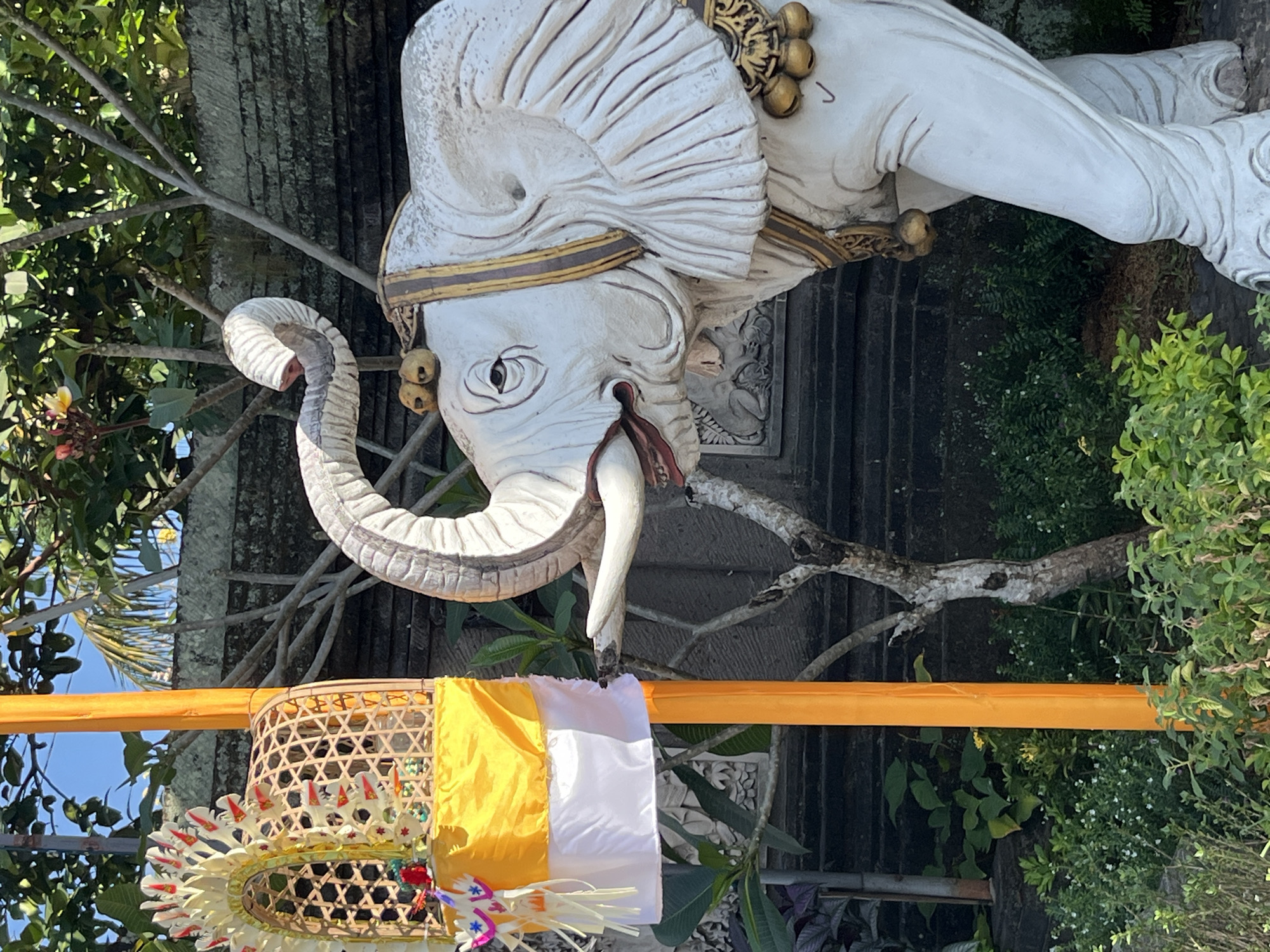 An outdoor, eye-level, close-up shot features a statue of a white elephant with ornate gold trim and bells on its ears and neck. The elephant's trunk is raised, and its mouth is slightly open, revealing red and white details inside. To the left of the elephant, a tall, bright orange pole stands vertically, adorned with a woven basket structure decorated with yellow and white fabric and colorful streamers. The background consists of lush green foliage, a stone wall, and a tree with bare branches. The scene appears to be in a tropical or subtropical environment.