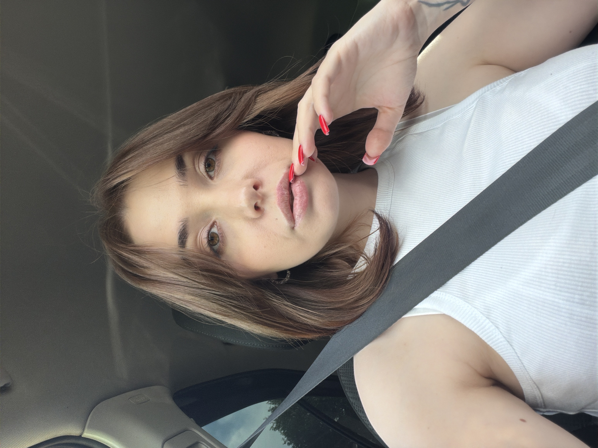 The media file is a vertical, close-up photograph of a young woman taken from the chest up, inside a car. She has medium-length, brown hair that frames her face, and her left hand is raised, with bright red fingernails, touching her lips. She is wearing a white, ribbed tank top and a black seatbelt is diagonally across her chest. Her expression is ambiguous, with her eyes looking directly at the camera and her lips slightly parted. The lighting suggests it is daytime, possibly overcast, given the diffused light and shadows. The interior of the car is visible in the background, with parts of the dashboard and ceiling. No text is visible in the image.