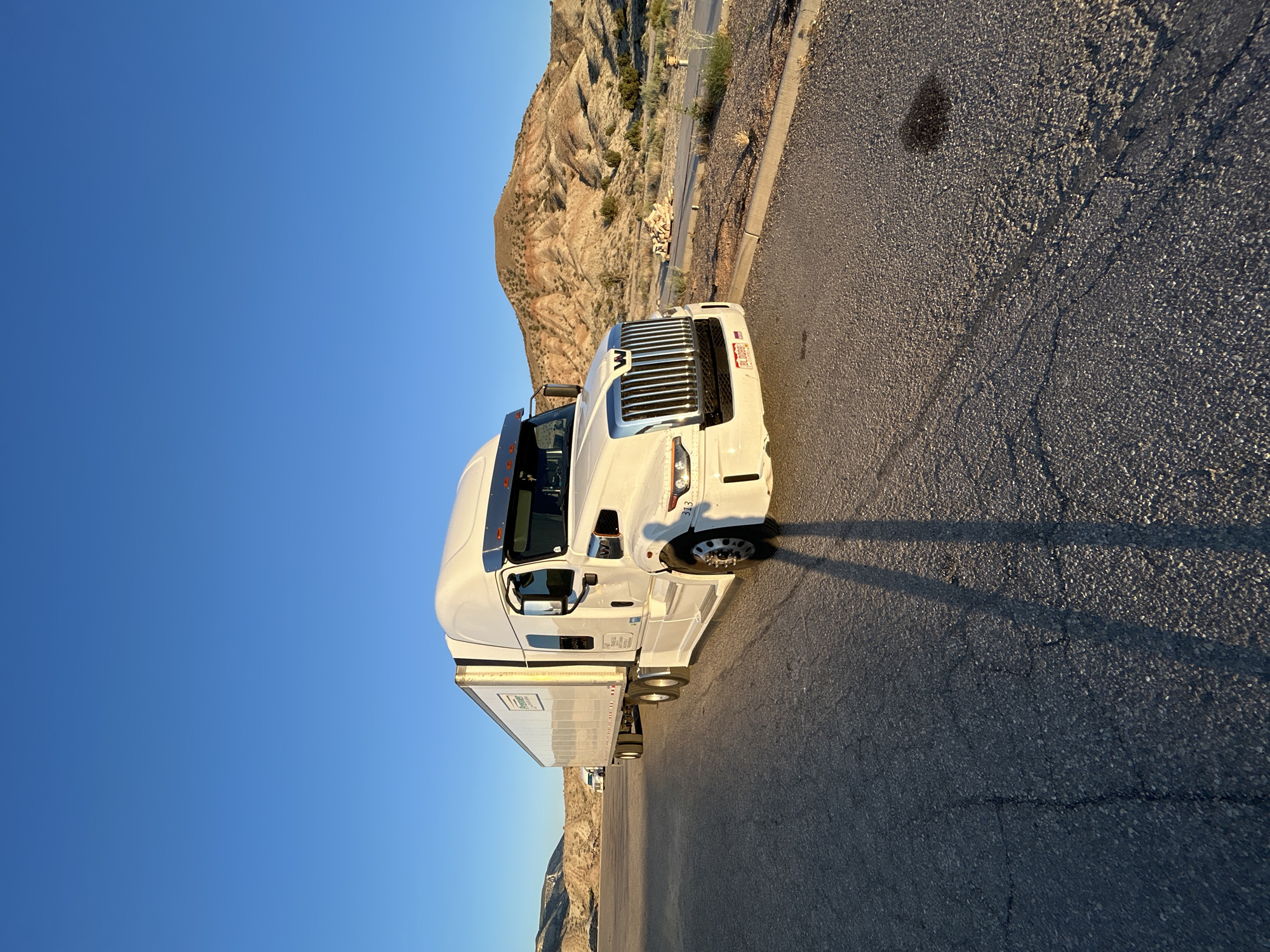 A white semi-trailer truck is parked on a cracked asphalt lot in a desert landscape. The truck has a long white trailer with "Premier" printed on the side in black lettering. The truck cab is white with a chrome grille and the Western Star logo. The sun is low in the sky, casting long shadows from the truck across the pavement. In the background, there are rolling hills with sparse vegetation and a clear blue sky. A second, smaller white truck is visible in the distance. The scene appears to be a rest stop or parking area in a rural location. There are no people visible in the image. The weather appears to be clear and sunny, likely in the late afternoon or early morning due to the long shadows and warm light.