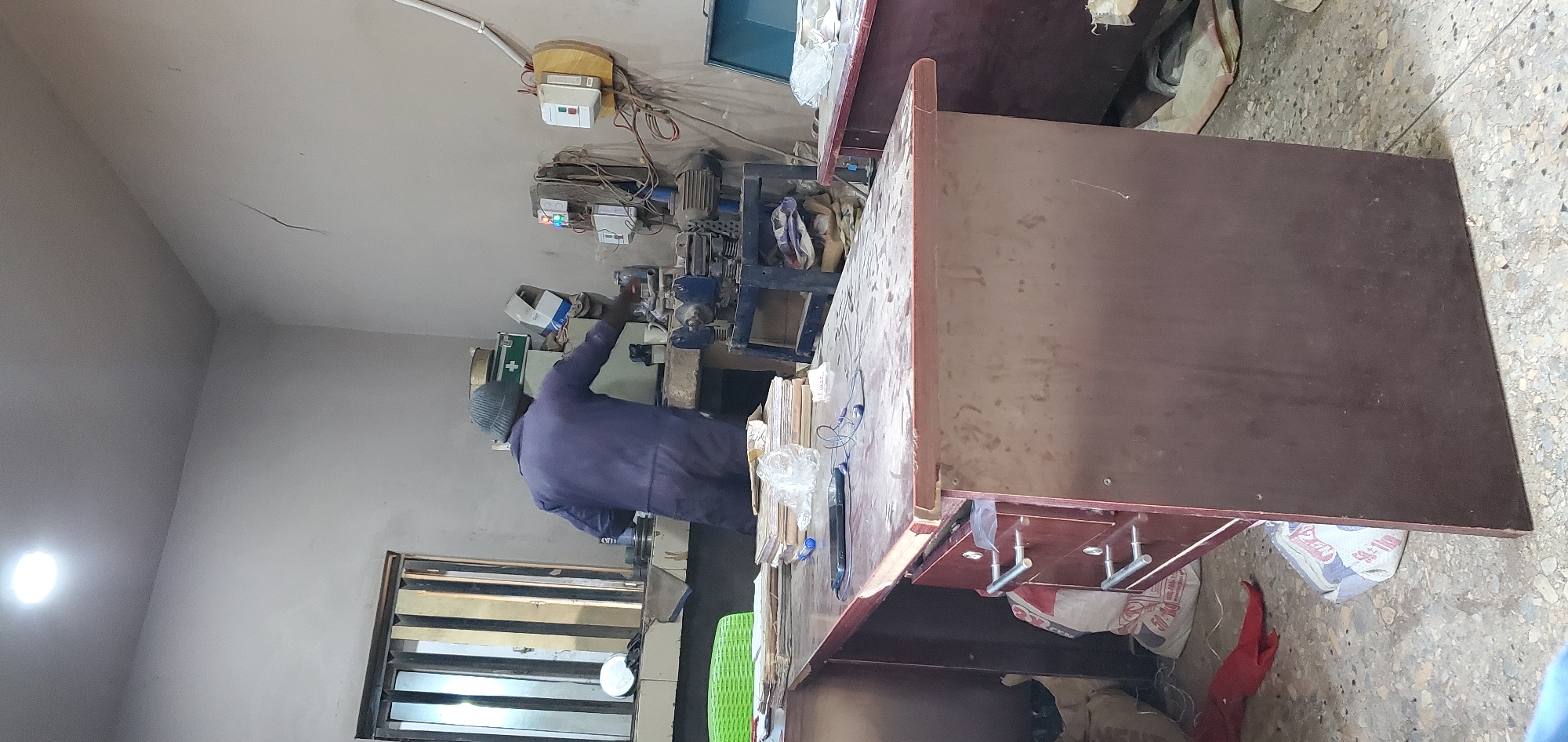 In Gura Top, Nigeria, a person wearing a dark blue jumpsuit and a green beanie is working in a cluttered workshop. The individual is standing behind a large wooden desk with drawers and is interacting with a piece of machinery on a workbench. Several stacks of papers and books are on the desk, along with a mobile phone and earbuds. In the background, there are more pieces of equipment, including a blue metal machine with an electrical panel. The walls are plain and somewhat cracked, and the floor is concrete with scattered debris. A window with vertical blinds is visible on the left. The overall impression is one of a functional but untidy workspace.