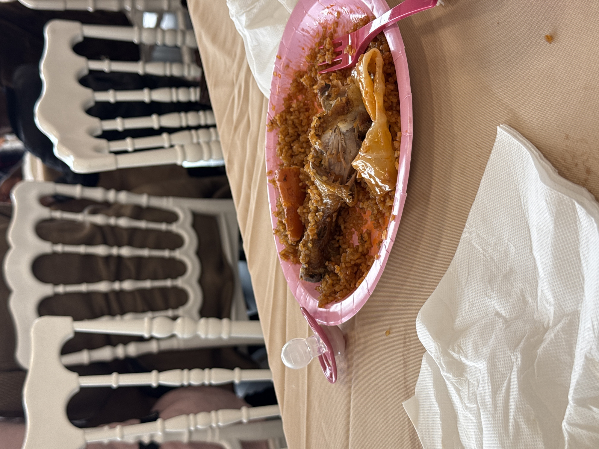 The image shows a pink disposable plate containing food, possibly bulgur with meat and vegetables, on a beige tablecloth. A pink plastic fork rests on the plate. To the right of the plate is a crumpled white paper napkin. A clear pacifier is attached to the bottom edge of the plate. The scene is set on a table in Domont, France. Chairs are visible in the background. The composition indicates an indoor environment with people present, likely during a meal. The image suggests a casual dining setting with food on a disposable plate.