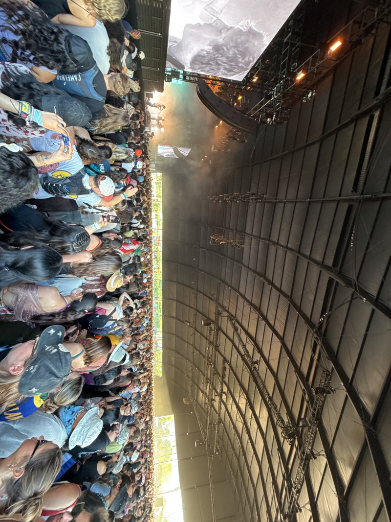 The image depicts a crowded music concert in Indio, California, United States. A large crowd is visible, facing a stage with video screens and concert lighting. A large tent-like structure covers the audience area. Some individuals in the crowd are holding up their phones, apparently recording the performance. The overall scene suggests a large-scale music festival or concert event.