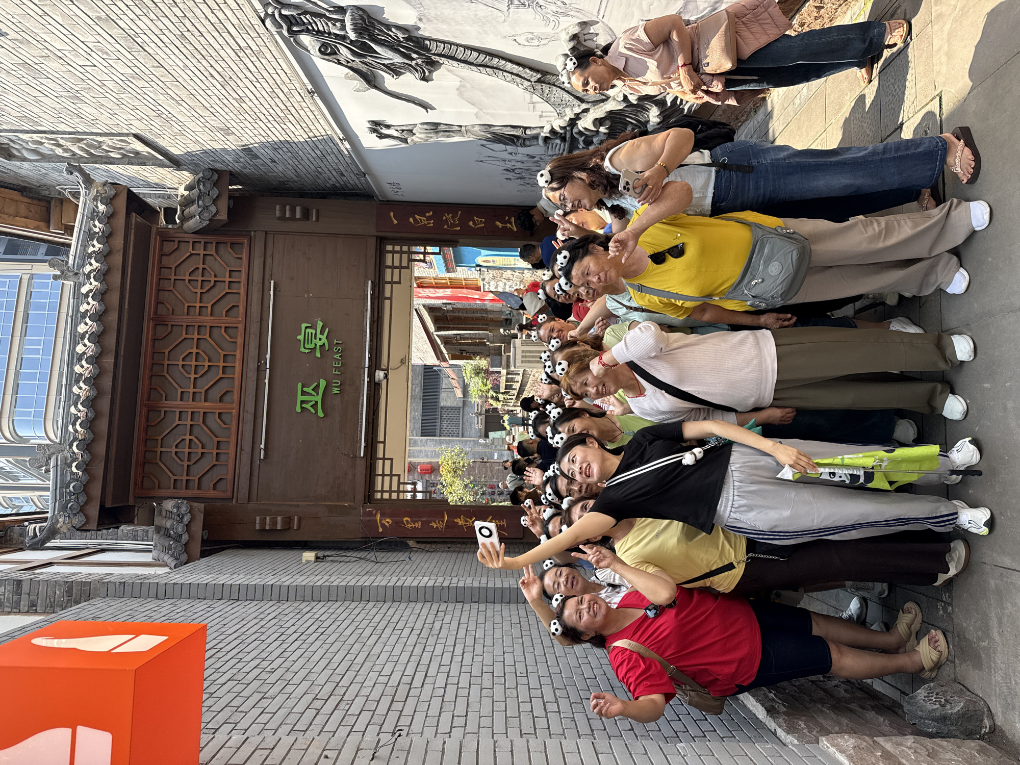 A group of approximately 15 to 20 women are gathered outdoors, posing for a photograph or selfie. Many of the women, particularly those in the front rows, are wearing small, black and white panda-themed hair clips or headbands. One woman in the foreground is holding a smartphone up, seemingly capturing a group selfie, while others are smiling, making peace signs, and looking towards the camera. Their expressions are cheerful and engaged, suggesting a happy and lively tourist experience.
The setting features traditional Chinese architecture, including a building with dark wooden facades, intricate lattice-work windows, and a dark tiled roof. Adjacent to this building, a large, ornate wall mural or relief sculpture of a dragon is visible, adding to the cultural aesthetic of the scene. The ground is paved with brick-like tiles. In the immediate foreground, a bright orange, square-shaped object with white markings is partially visible.
The scene is brightly lit by natural daylight, indicating midday or early afternoon, with clear shadows cast by the buildings and people. The women are dressed in casual touring attire, with some wearing sunglasses or carrying crossbody bags. One woman is holding a green and yellow collapsible umbrella.
Visible text on a sign above the building entrance reads "WÚ FEAST" in green English letters, accompanied by Chinese characters, likely "吾宴". The overall atmosphere suggests a group of tourists enjoying a cultural site in Yuzhong District, China, an impression reinforced by the traditional architecture and panda accessories.