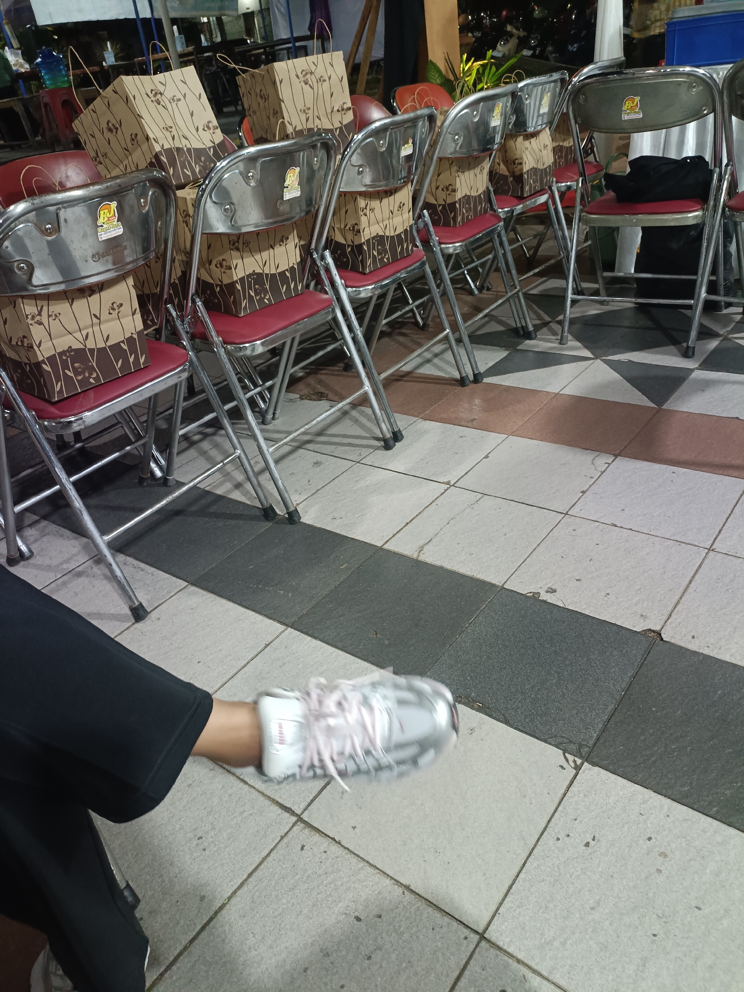The image shows a row of red and silver folding chairs with brown paper bags on them, likely staged for an event. The chairs are arranged in a tiled area that appears to be outdoors, possibly a public space or a venue in Condongcatur, Indonesia. The foreground features a person's leg and foot wearing a white sneaker with pink laces, indicating their presence at the scene. The background is blurred, but suggests a nighttime setting with ambient lighting.