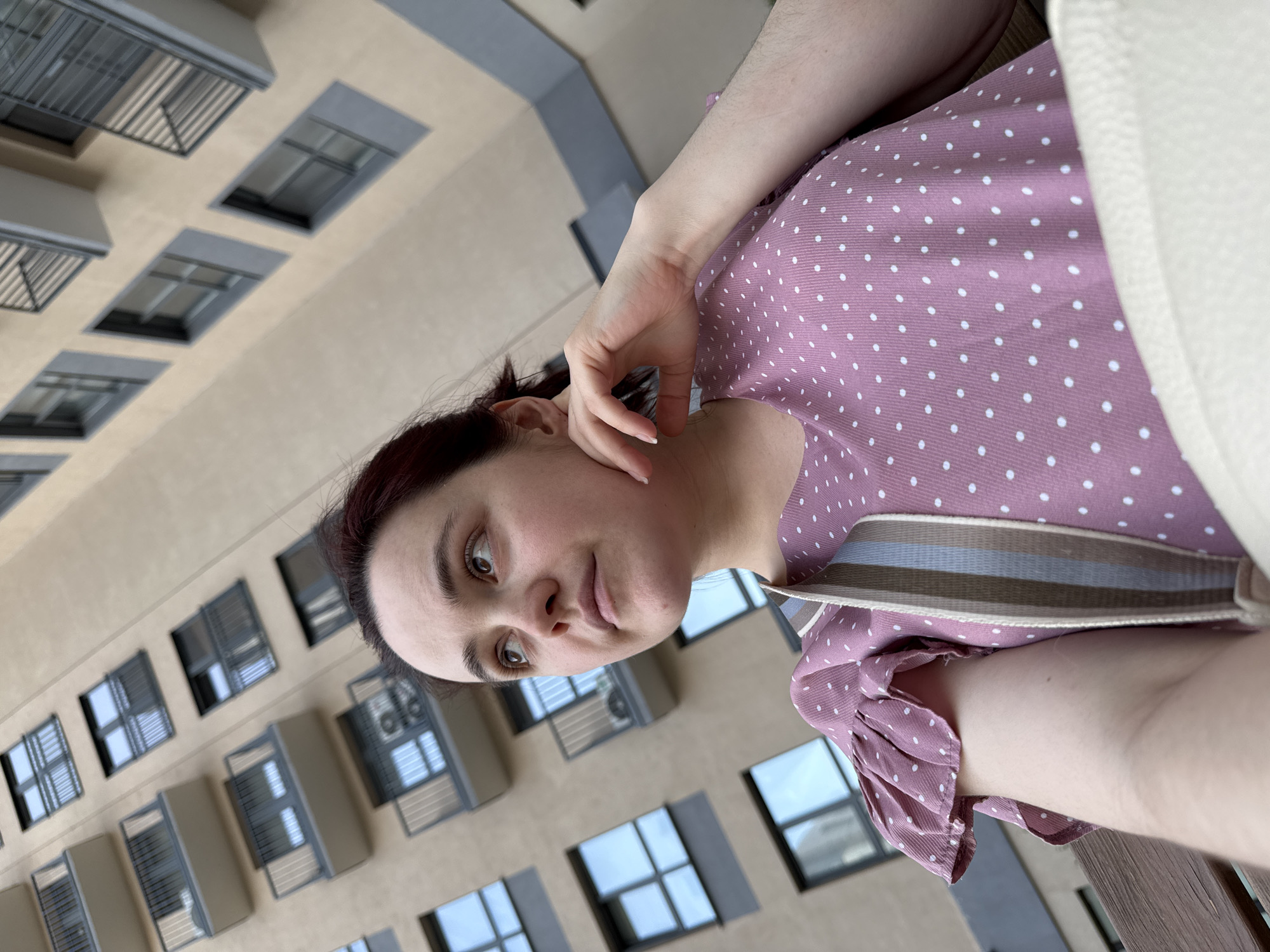 A person wearing a pink and white polka dot shirt is holding their hand to their face, looking at the camera. They are standing in front of a multi-story building with windows and balconies, potentially in an urban setting, though specific location identification in Izhevsk, Russia cannot be confirmed without additional information.