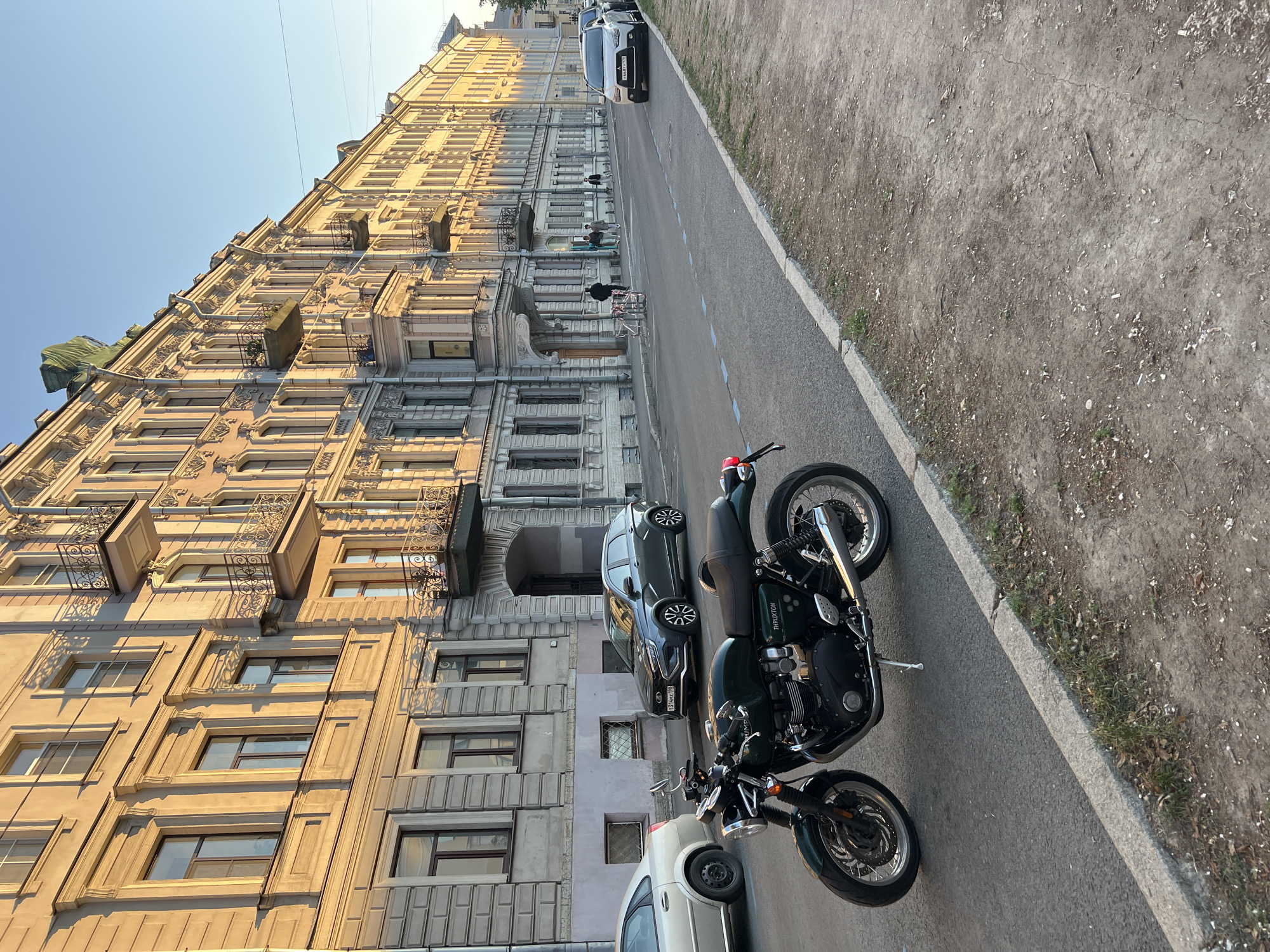The image depicts a street scene in Saint Petersburg, Russia, likely in the late afternoon, as indicated by the warm, long shadows cast by the buildings. The sun is low in the sky, illuminating the ornate facades of a historic apartment building on the left. The building features a mix of architectural styles, with cream-colored lower floors and lighter, almost white upper floors, adorned with intricate detailing, balconies, and numerous windows.
On the street, a dark green motorcycle is parked. It appears to be a Triumph Thruxton, with its distinctive styling and chrome accents prominently displayed. The motorcycle is positioned on the asphalt road, which is bordered by a rough, dirt and gravel verge with sparse greenery. Further down the street, several cars are parked, including a dark SUV and a lighter sedan. A few pedestrians are visible walking along the sidewalk, suggesting a normal day in the city. The sky is clear and blue. The overall impression is one of urban tranquility bathed in the golden light of a setting sun. No discernible text is visible on the buildings or the motorcycle, though a license plate is present on the dark SUV.