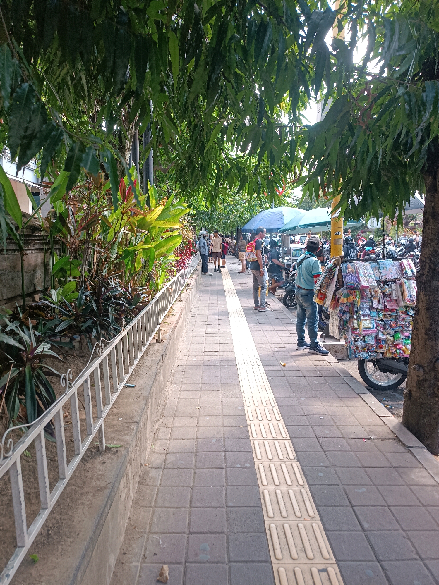 The image depicts a pedestrian sidewalk lined with trees and lush plants. On the right side of the sidewalk, a vendor displays an assortment of colorful trinkets and toys mounted on a cart, which is attached to a motorcycle. Several people are seen walking along the sidewalk and near the vendor. In the background, a row of motorcycles is parked, suggesting a busy urban area. The sidewalk itself is paved with square tiles and features a tactile paving strip for visually impaired pedestrians. The presence of the plants and the architecture suggests this scene is likely in Tabanan, Indonesia. The overall impression is a typical street scene in a bustling Indonesian city.