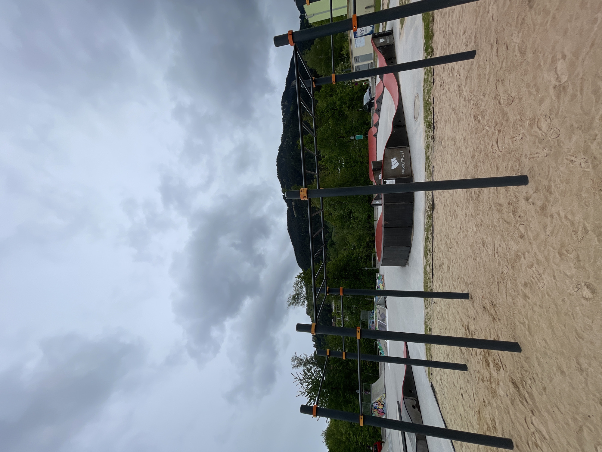 The image shows an outdoor calisthenics park, featuring metal bars and ladders set against a backdrop of a mountain and a cloudy sky. The ground is covered in sand. Equipment with "PARKITECT" branding can be seen. The location appears to be in a city, but the specific city and region are not identifiable. There are no people visible in the frame.