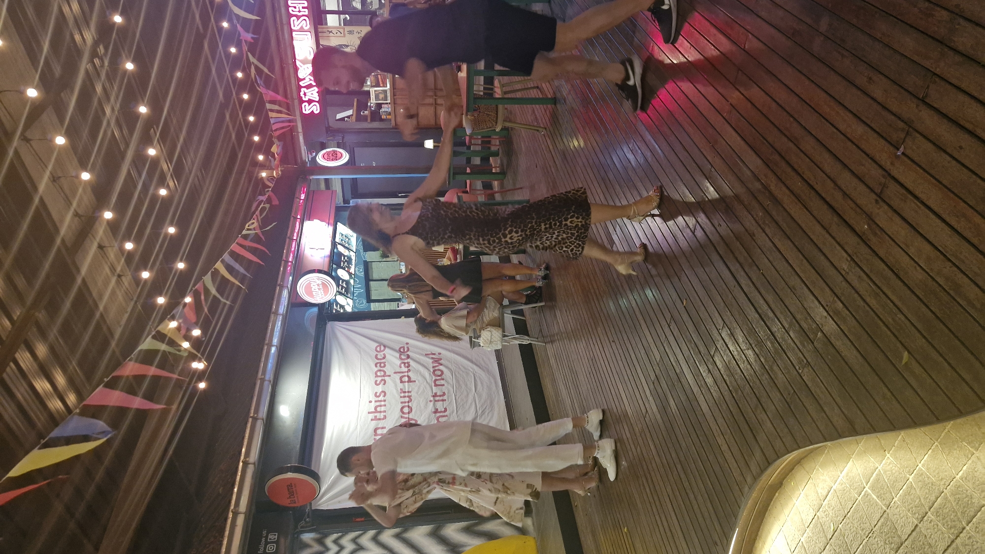 The image depicts an outdoor venue with a wooden floor, possibly a patio or deck, illuminated by strings of lights. Several people are present, with a notable group engaged in dancing. In the foreground, a couple in white attire is dancing, with the man in a white shirt and pants and the woman in a floral dress. To their right, another couple is dancing; the woman wears a leopard-print dress and high heels, while the man is in a dark t-shirt and shorts. Behind them, other individuals are seen sitting and standing, observing the scene or engaged in conversation. The background features storefronts with signage, including "la barra.", "HAPPY", and "SAXY SUSHI," suggesting a commercial area with food and entertainment establishments. The presence of colorful triangular flags strung across the ceiling adds to the festive atmosphere. The overall scene suggests a lively evening in a public gathering space. The location is identified as Ban Bang Rak, Thailand.