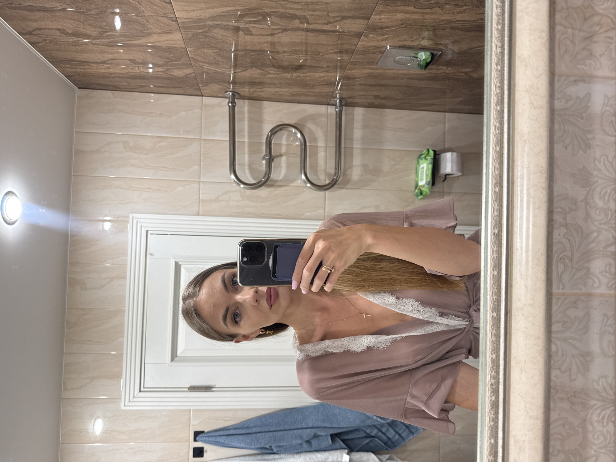 In a bathroom, a woman with long blonde hair takes a mirror selfie. She is wearing a light pink robe with white lace trim and a gold necklace with a cross pendant. She is also wearing earrings and rings. The bathroom has beige and brown tiled walls and a silver towel rack. There is a white door in the background and two towels hanging on the left side. The scene appears to be indoors.