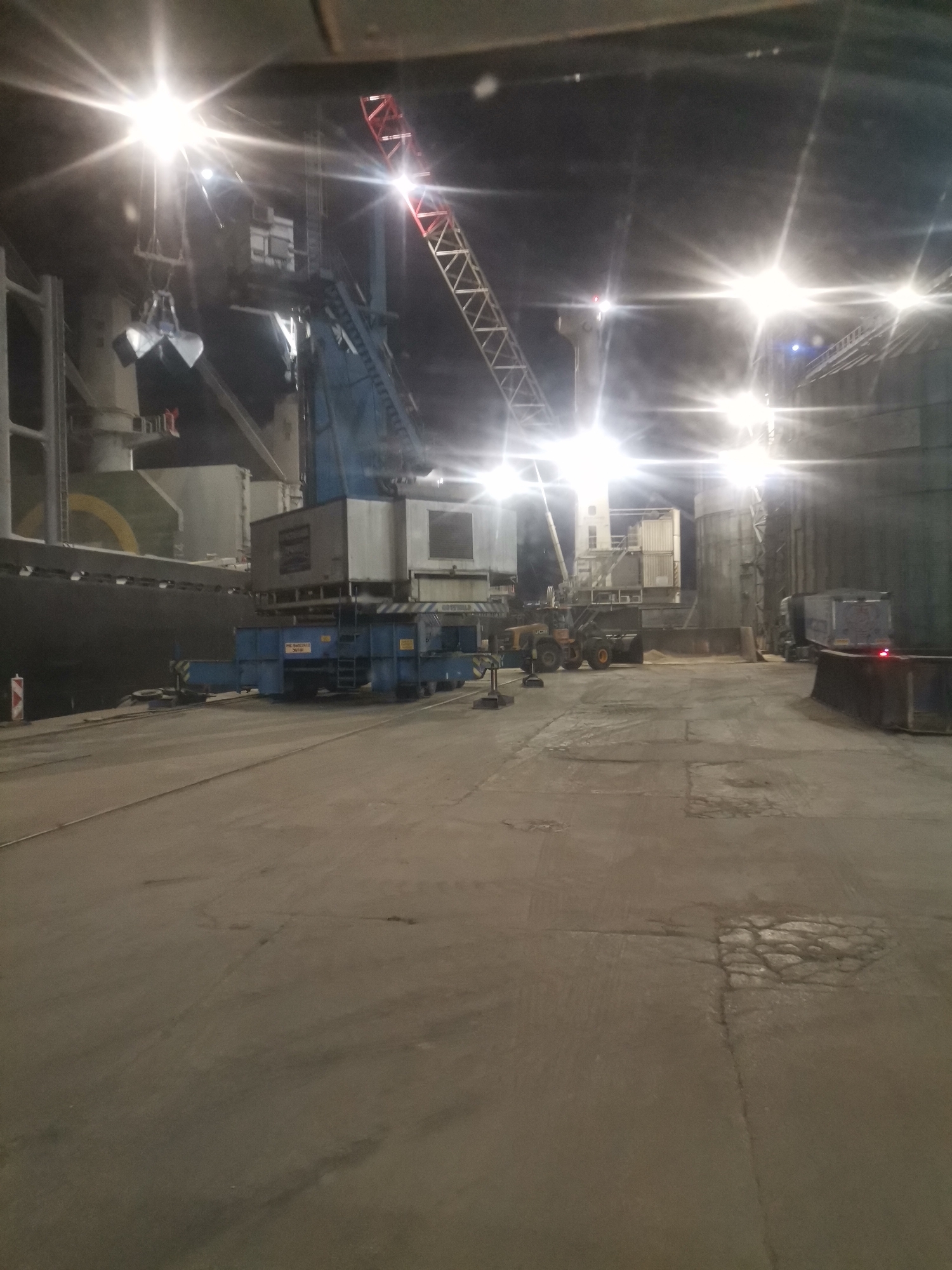 The image depicts a harbor scene at night. Several bright overhead lights illuminate the area. In the foreground, there is a large concrete surface with cracks and patches. Further into the scene, a ship is docked alongside a pier. A large blue crane is situated on the pier, and other industrial equipment is visible, including a front-end loader. In the background, large storage containers are seen. The location is likely within the port area of Varna, Bulgaria, a major port city. There are no people visible.