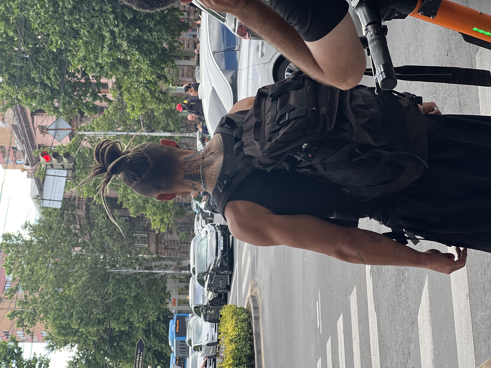 The image captures a person from behind, standing on a city street in Yerevan, Armenia, during what appears to be midday or early afternoon under clear skies. The primary subject has dark dreadlocks styled into an updo and features extensive tattoos with tribal-like patterns on their neck and right arm. They are dressed in a black sleeveless top and a black tactical backpack with multiple straps and compartments. A silver chain is visible around their neck, and small hoop earrings are also discernible.
To the right, the arm of another individual is visible, holding an orange and black e-scooter. The scene depicts a typical urban street with a pedestrian crosswalk marked on the asphalt. A red traffic light is illuminated on the left side of the frame. A line of parked cars extends into the background, including a black vehicle with an Armenian license plate "50 RU 505." Further down, a blue public bus is partially visible.
The street is lined with green trees and buildings of varying architecture, some featuring reddish-brown facades. A street sign with Armenian text "ԱՄԻՐՅԱՆ փ" (AMIRYAN str.) confirms the location as Amiryan Street. A low, manicured hedge of green bushes runs along the curb. The overall impression is a vibrant city environment.