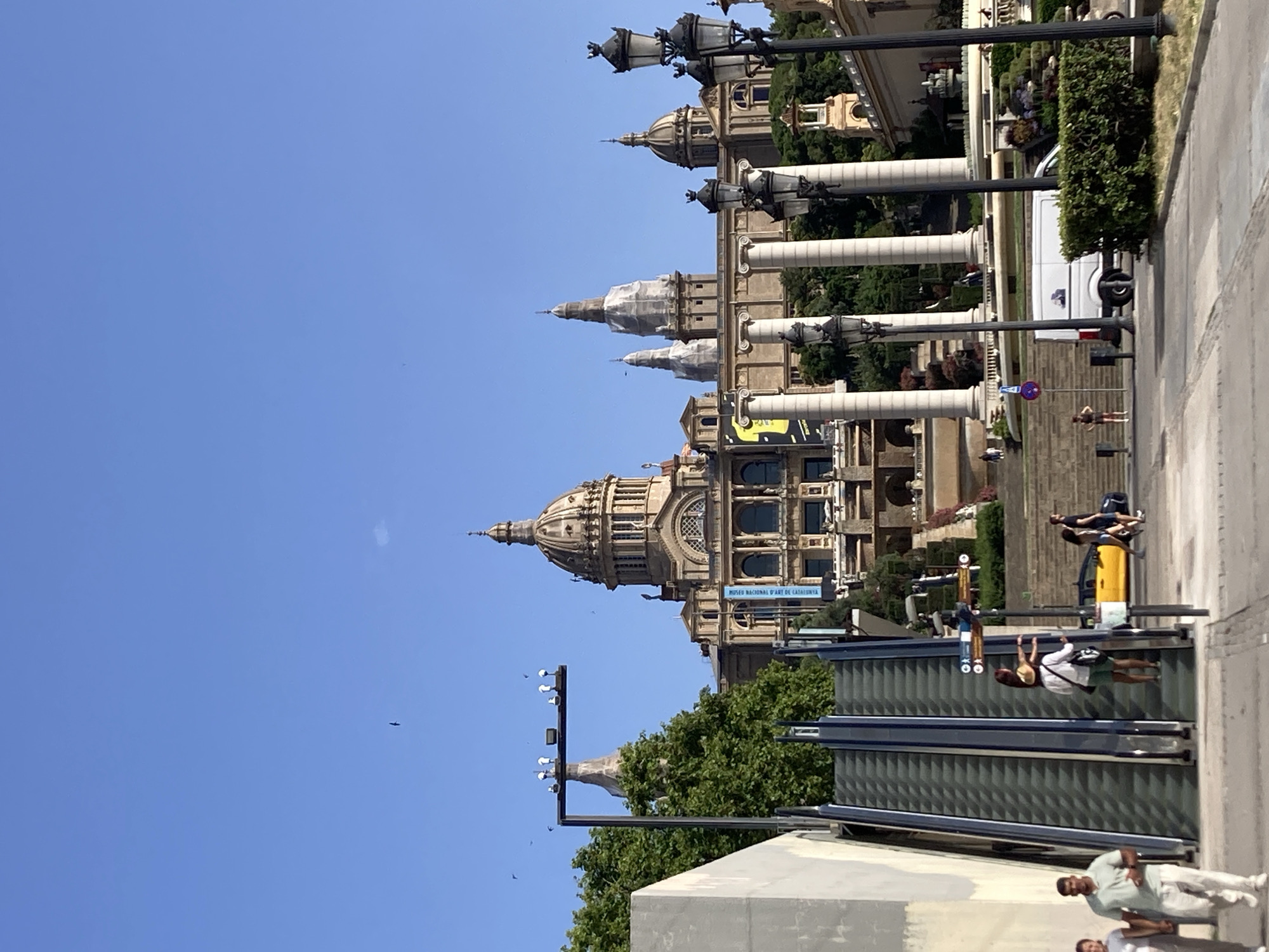 The image captures a sunny day in Barcelona, Spain, likely in the afternoon given the bright light and clear blue sky. The scene is dominated by the imposing architecture of the Museu Nacional d'Art de Catalunya, a grand building with a distinctive dome and multiple spires. The building is a mix of classical and baroque styles, with large columns and ornate detailing.
In the foreground, there are several people engaged in various activities. One person is walking up a set of stairs, while others are seated or standing near the entrance of a modern structure with reflective surfaces, possibly an escalator or covered walkway. A white van is parked on the side, partially obscured by lush green trees and bushes. The overall atmosphere appears relaxed and tourist-oriented.
There are several notable details:
- A blue sign on the museum building reads "MUSEU NACIONAL D'ART DE CATALUNYA," confirming the location.
- Several lampposts with intricate designs are visible, suggesting a well-maintained public space.
- There are a few birds flying in the sky.
- The weather is clearly sunny and warm, with no clouds present.
The image is oriented vertically, suggesting it might have been taken with a phone camera held in portrait mode. The perspective looks upwards towards the museum, highlighting its scale and grandeur against the clear sky.