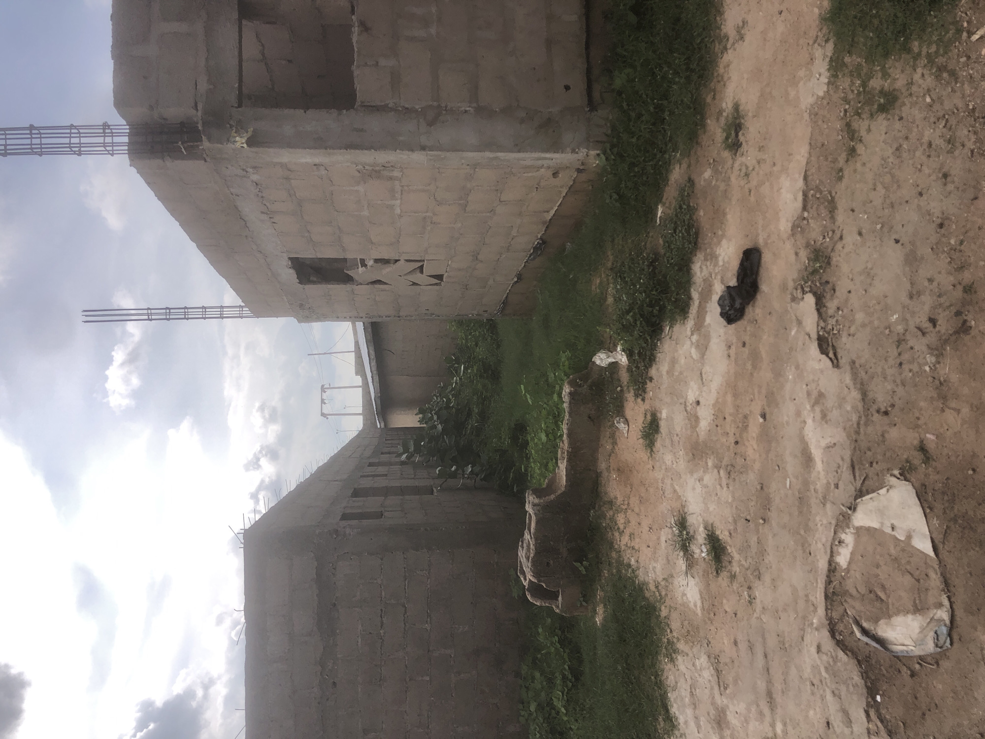 The image depicts an outdoor scene in Jalingo, Nigeria, focusing on two unfinished concrete block buildings. The buildings are in a state of construction, with exposed rebar at the top of one structure and open window frames. The ground in the foreground is a mixture of dirt and sparse grass. In the middle ground, a weathered, stone-like object of indeterminate purpose rests on the grass. A pair of dark shoes is visible on the dirt path towards the right side of the image. Vegetation, including tall grass and bushes, surrounds the buildings. The sky above is partly cloudy, with patches of sunlight breaking through. The overall impression is one of an ongoing construction project in a semi-urban or rural setting.