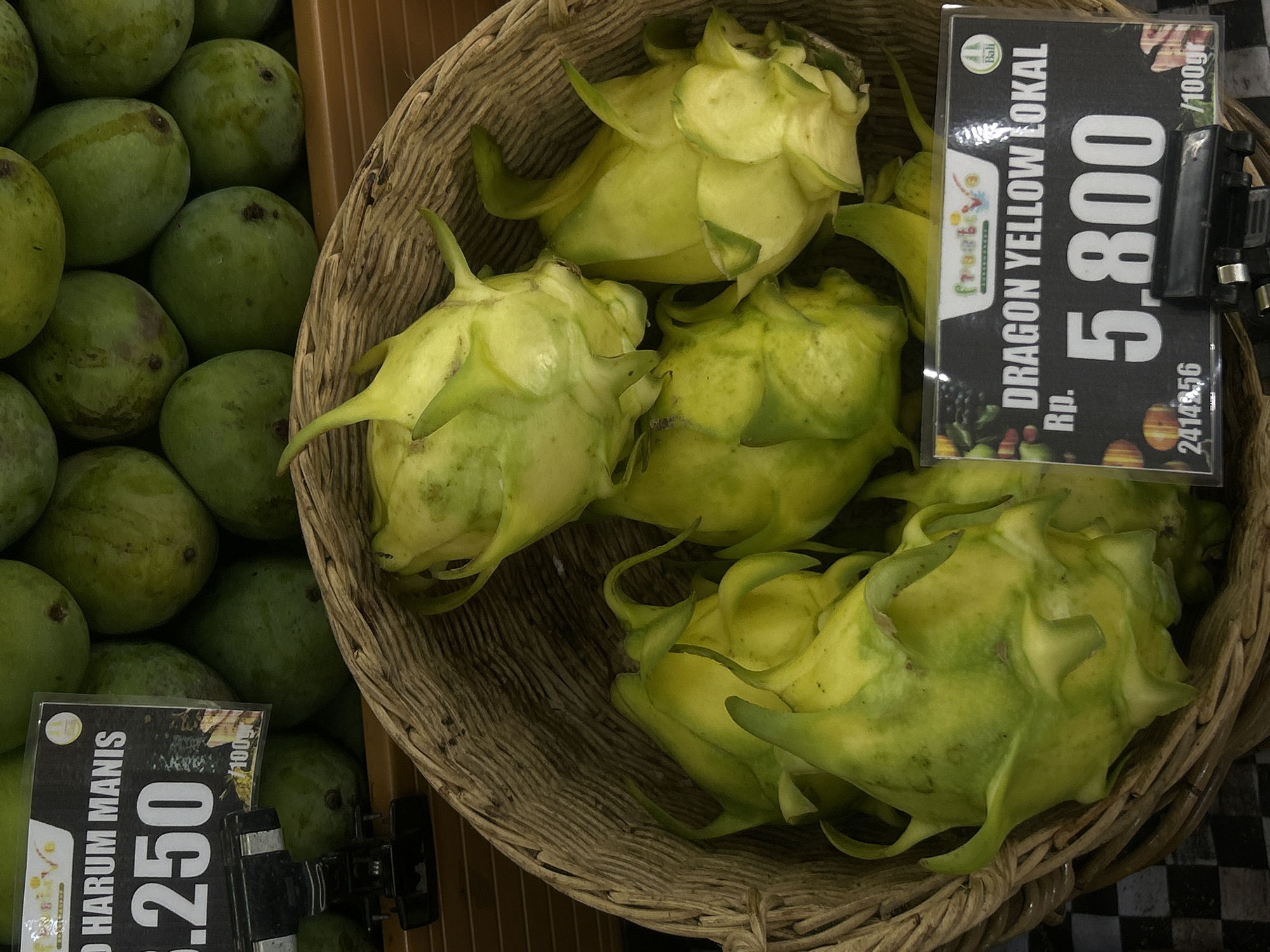 The photograph depicts a display of fruit within a retail setting, likely a grocery store or market in North Kuta, Indonesia. On the left, green mangoes are arranged in a pile. Adjacent to the mangoes, a woven basket contains several yellow dragon fruits. Price tags are attached to the displays, providing information about the cost per 100 grams. The price tag for the mangoes reads "HARUM MANIS 6.250", while the dragon fruit is labeled "DRAGON YELLOW LOKAL Rp. 5,800". The price tag for the dragon fruit also includes the logo of "frostive" and the word "Bali".