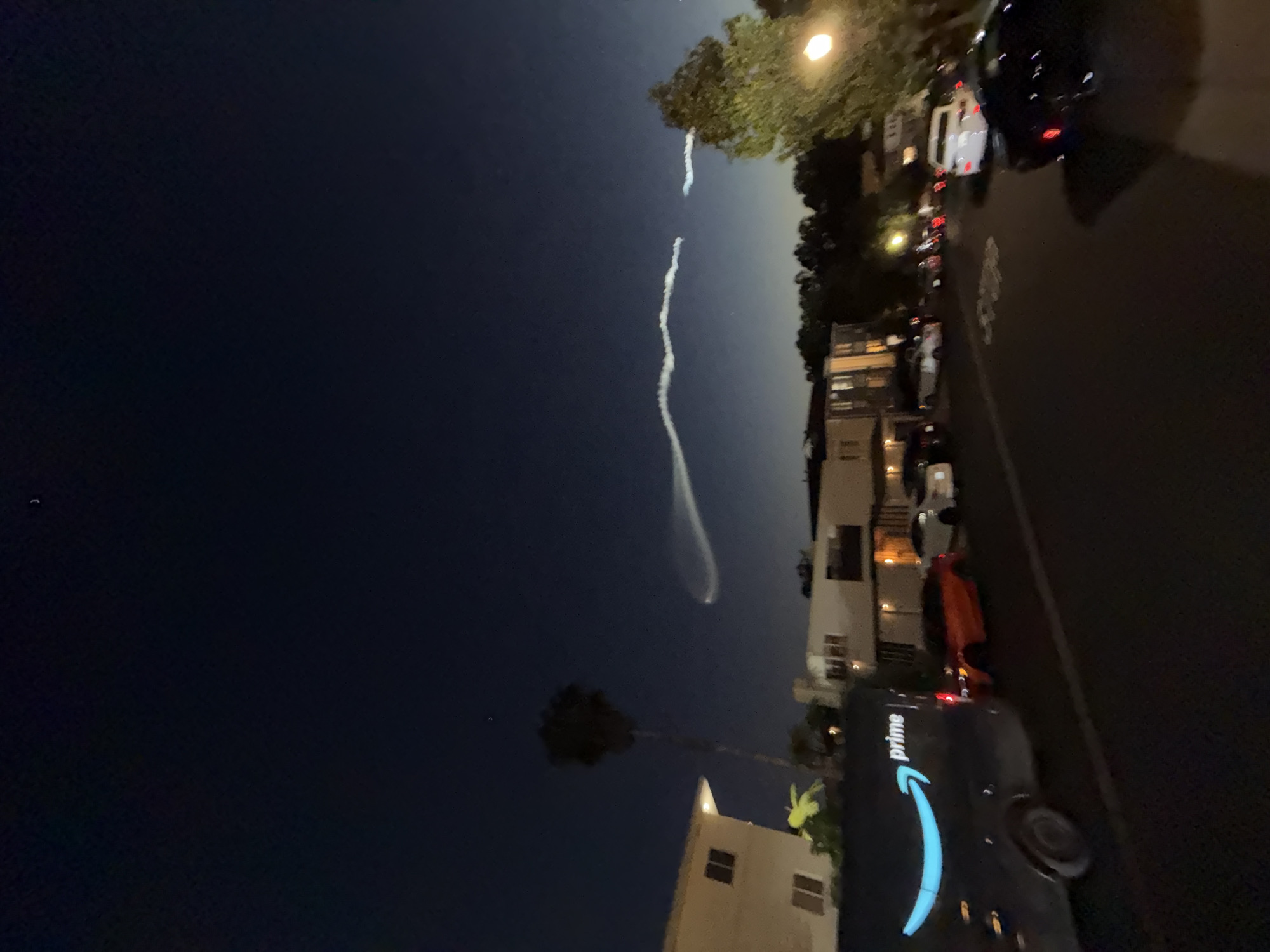 The image captures a night scene in Los Angeles, United States of America. In the sky, a distinct contrail is visible, suggesting the recent passage of a rocket or other high-altitude aircraft. The contrail has a curving shape and dissipates as it rises.
On the ground, a residential street lined with houses and parked vehicles is seen. An Amazon Prime delivery van is parked on the right side of the street. The street is illuminated by streetlights, and the houses show various levels of lighting. Trees are interspersed among the buildings.
