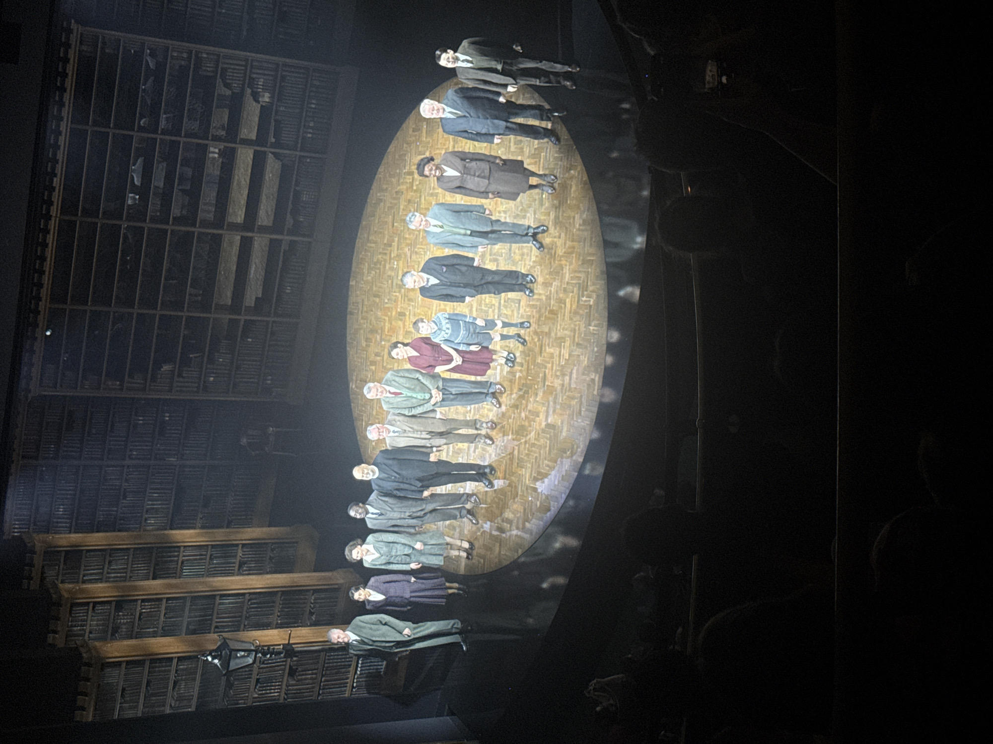 The image shows a scene from the stage production of "Harry Potter and the Cursed Child" at the Palace Theatre in London, United Kingdom. The set depicts a scene from a library or historical archive, featuring rows of shelves filled with what appear to be books and documents. In the foreground, actors in period-appropriate costumes are lying on a large, oval-shaped platform with a parquet floor pattern. The actors appear to be a mix of male and female, and their poses might represent a dream sequence or a metaphorical representation of time or ancestry. The photograph is taken from a seated audience perspective. The house is dark, but faces can be seen in the seats.