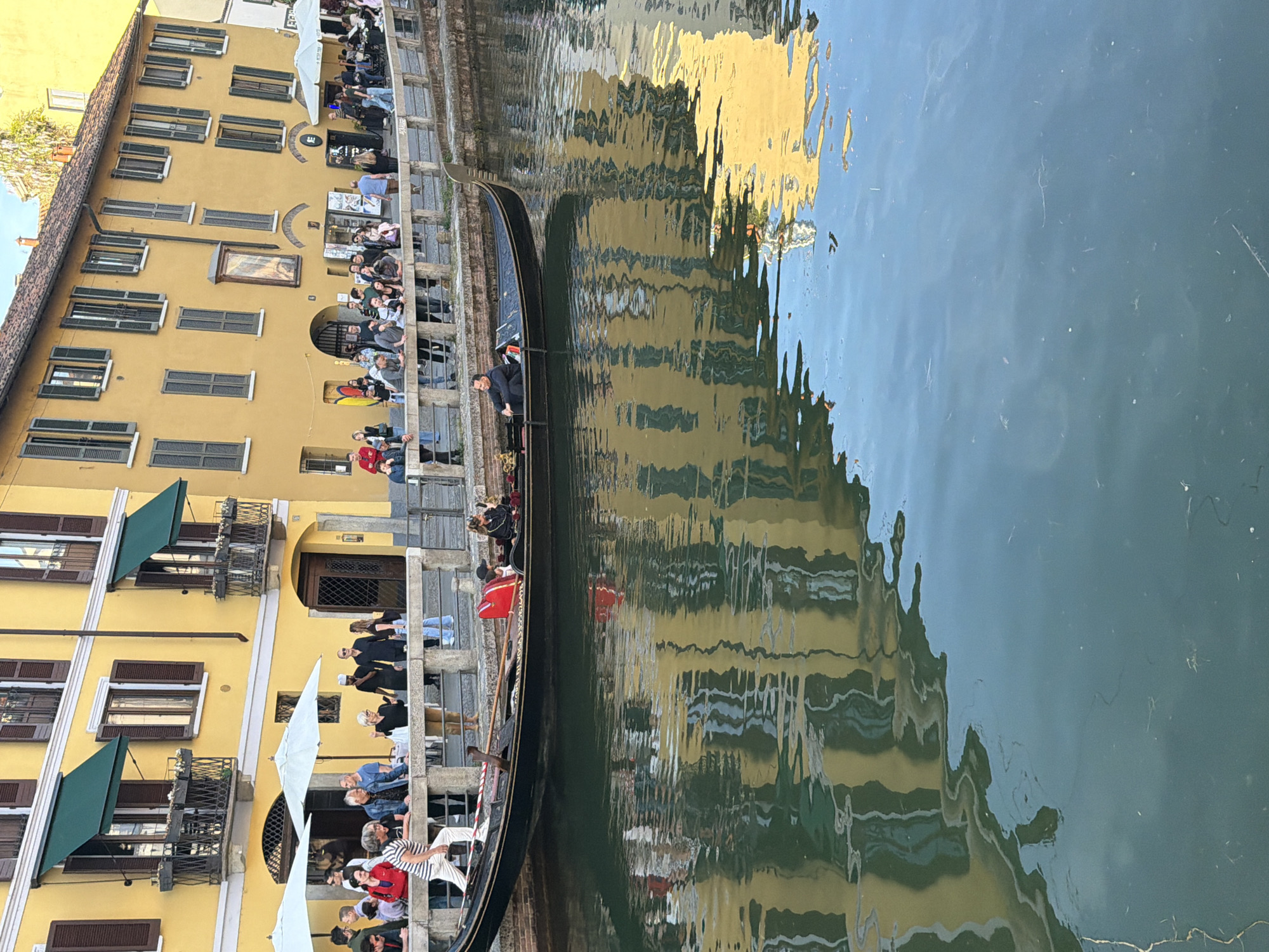 This image depicts a canal scene in Milan, Italy, likely in the Navigli district. A gondola carrying passengers is being navigated along the waterway. On the canal's edge, a yellow building with green shutters stands, with people seated or standing along the canal's side, possibly at restaurants or cafes. The building's reflection is visible in the canal's water. The scene appears to be during the daytime under relatively bright sunlight.