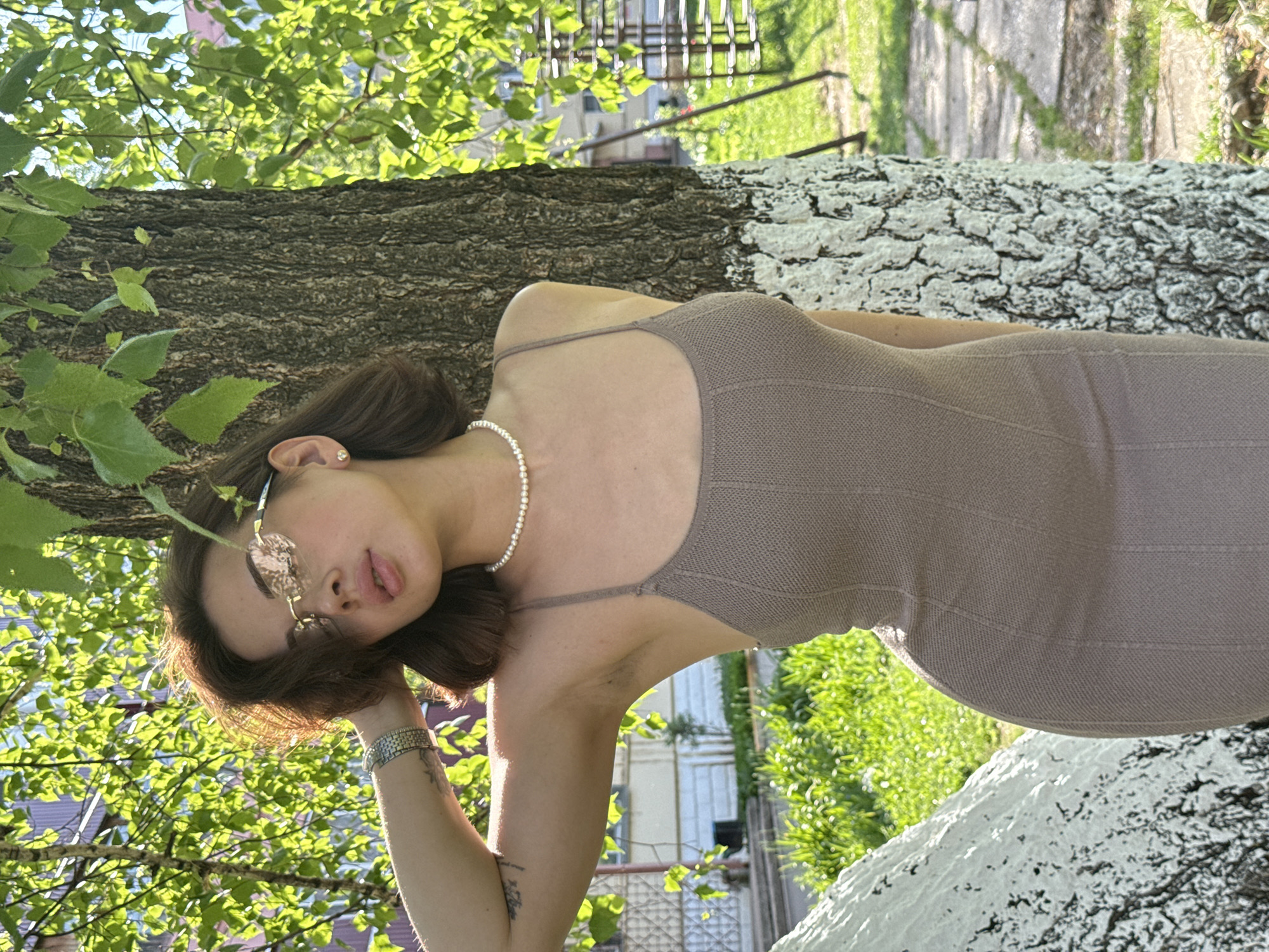 A young woman is posing for a photograph outdoors. She is wearing a brown dress, a pearl necklace, and sunglasses. Her right arm is bent and positioned near her head. She is standing next to a tree trunk that is partially painted white. Green leaves are visible in the background, along with other greenery. Beyond the trees, there are glimpses of buildings and structures. Based on visual cues such as architectural details and general appearance, the location may potentially be in Sarapul, Russia.