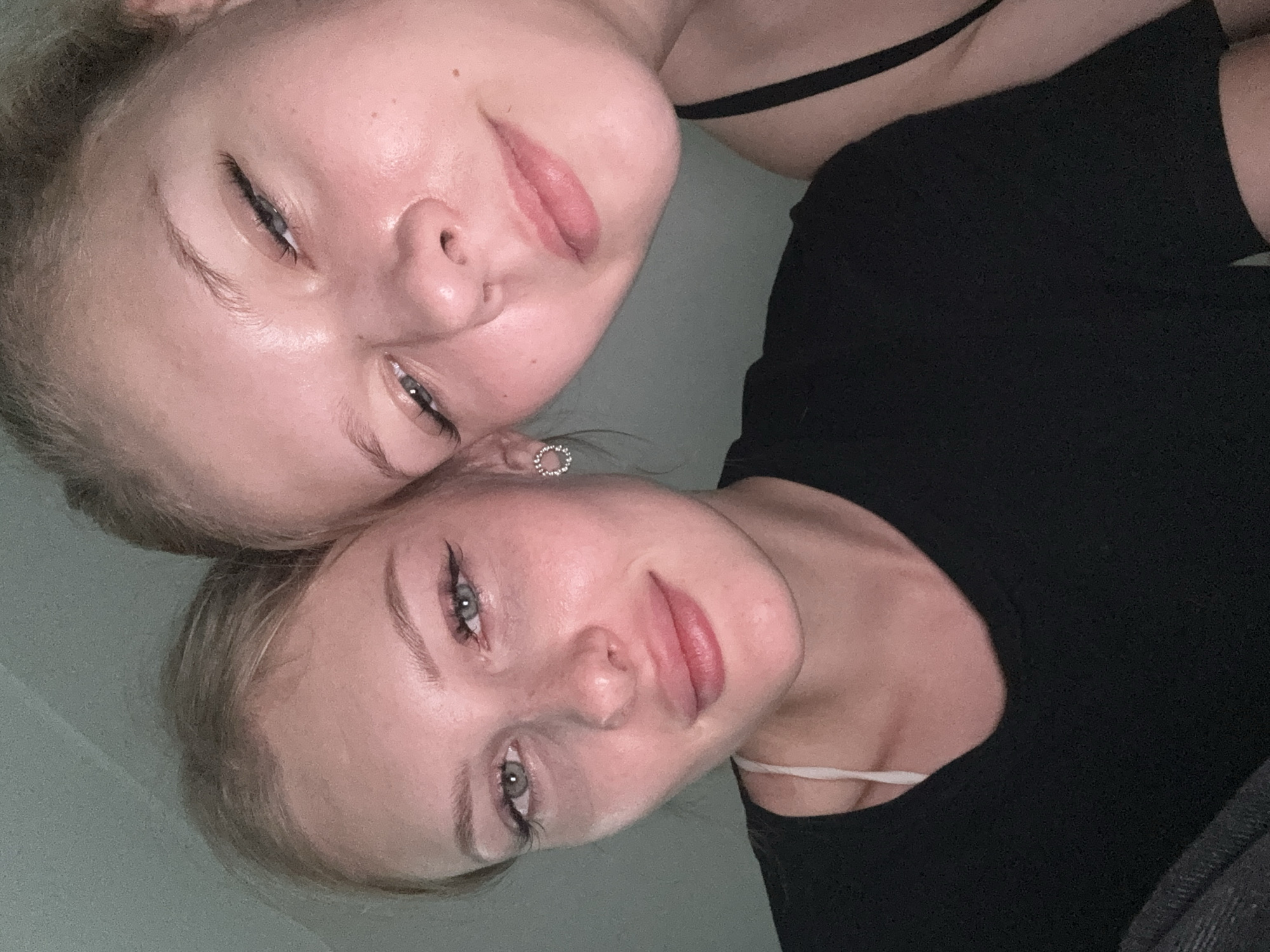 The image is a close-up, selfie-style photograph of two young women. The woman on the left is in focus, looking directly at the camera with a slight smile. She has blonde hair pulled back, blue-green eyes with visible eyeliner and false eyelashes, and light skin with freckles. She is wearing a black t-shirt with a white strap visible underneath. She has a small, circular, jeweled earring in her left ear.
The woman on the right is partially out of focus and to the right of the frame. Her face is angled away from the camera, and only her right eye and part of her forehead and cheek are clearly visible. She also appears to have blonde hair. She is wearing a black tank top.
The background is a plain, light green-grey wall. The lighting appears to be artificial and somewhat dim, casting a pinkish hue on the skin of both subjects. There are no visible landmarks or context clues to suggest a specific location within the city of Perm, Russia. The overall impression is that of a casual, personal photograph taken indoors.