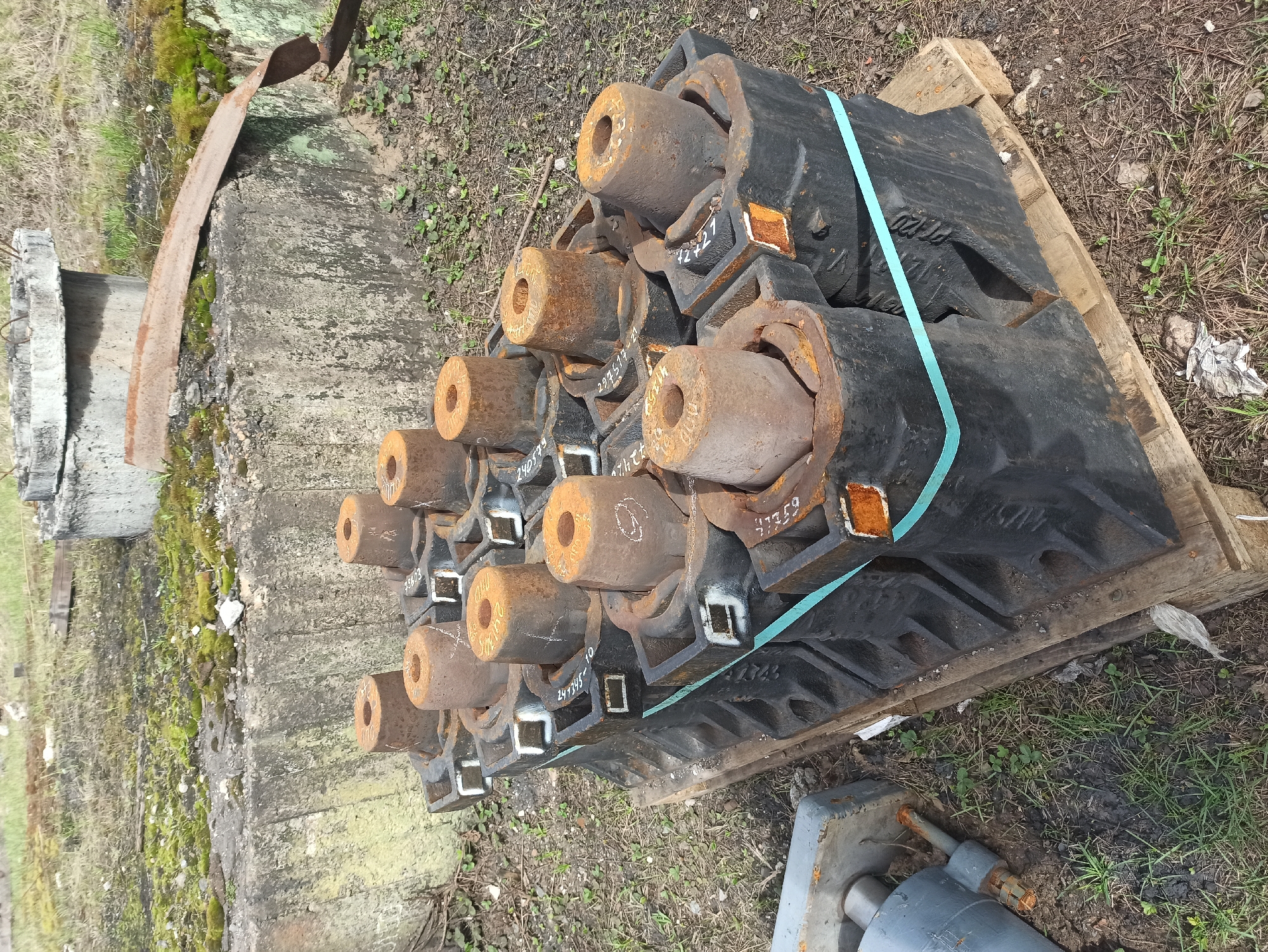 The image shows a collection of heavy, industrial components stacked on a wooden pallet in an outdoor environment. The components are primarily black and cylindrical, with rusted, hollow centers. Some have white markings, possibly serial numbers or identifiers. A blue plastic strap secures the components on the pallet. The pallet rests on the ground amid sparse vegetation and dirt. To the left is a weathered concrete structure covered in moss. A piece of rusty metal leans against the structure. In the foreground, to the right, is a grey metal object with protruding rods. The scene is located near Perm, Russia, as indicated by the marking “Perm” on one of the components.