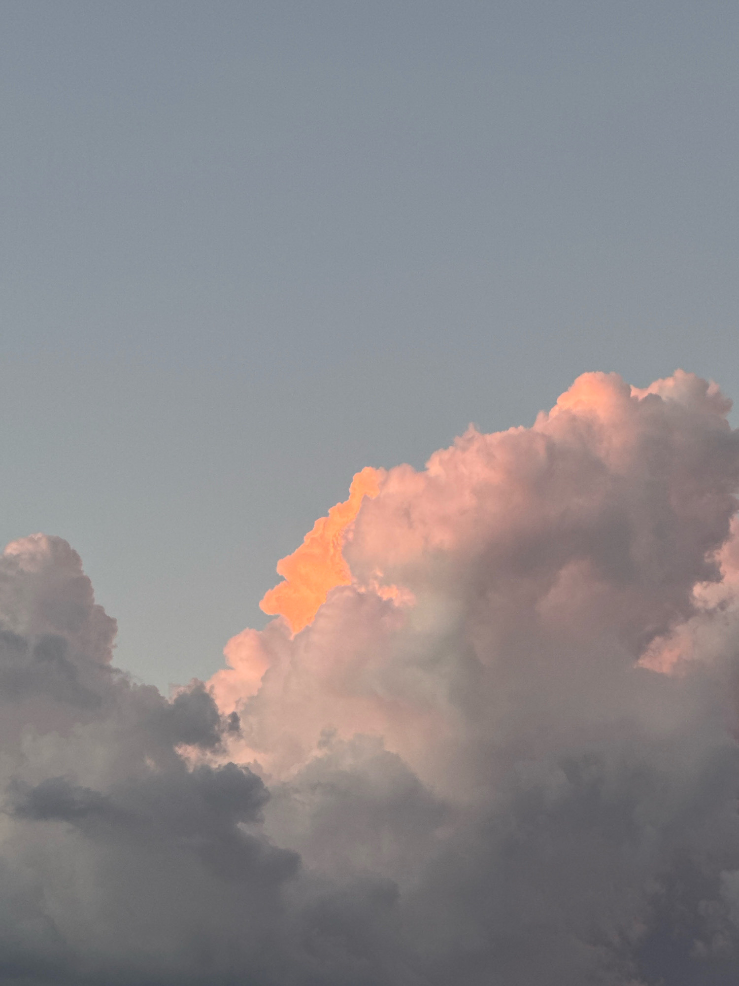This image depicts a sky filled with dramatic clouds, tinged with the warm hues of sunset or sunrise. The sky is a muted blue-grey, providing a stark contrast to the vibrant colors of the clouds. In the upper right quadrant, a large cumulus cloud is illuminated from behind by a bright, fiery orange light, giving it a glowing appearance. This central cloud is surrounded by other, darker clouds that appear to be catching the light from different angles, resulting in a mix of pink, grey, and white tones. The overall impression is one of a powerful and majestic natural display. The scene is not identifiable as being located in Capaci, Italy, as there are no terrestrial features visible in the image.