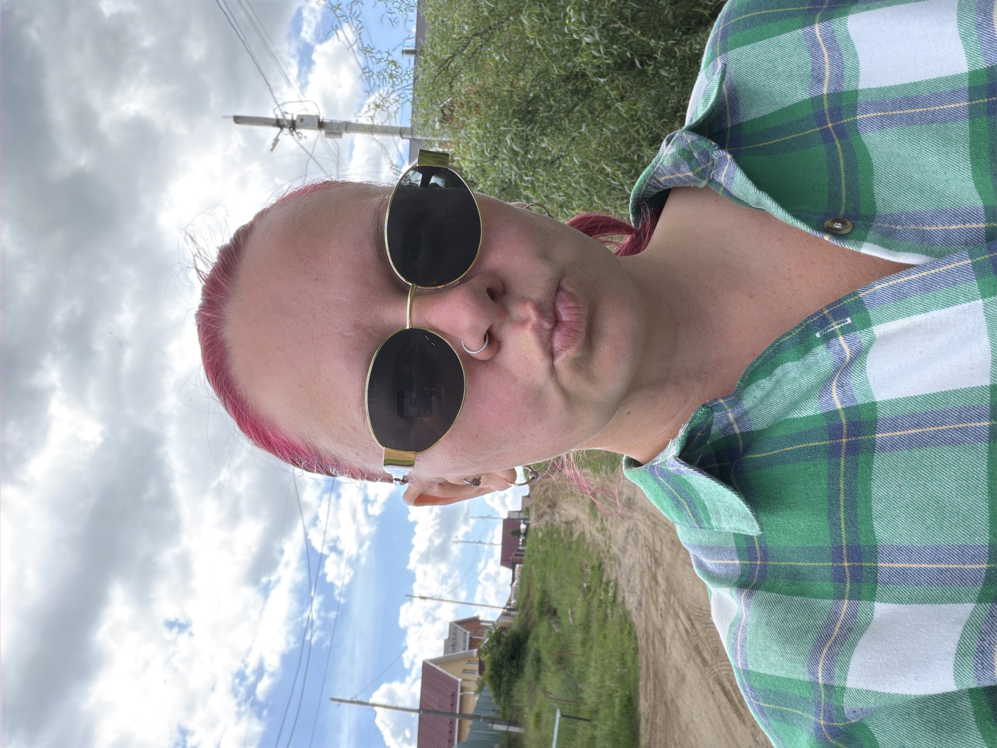 The image is an outdoor close-up portrait of an individual in Chaikovsky, Russia. The subject, of light complexion, has bright pink hair and is looking directly at the camera. The individual wears gold-framed, oval-shaped sunglasses with dark lenses, a silver hoop piercing in the right nostril, and multiple ear piercings, including at least two hoops on the left earlobe, a small hoop on the right earlobe, and other studs or barbells higher on the left ear. The lips are pursed in a pouting expression. The individual is wearing a long-sleeved, collared plaid shirt in shades of green, light blue/white, yellow, and dark blue, with the top buttons undone.
The background behind the individual features a rural or semi-rural residential landscape under a partly cloudy sky with visible blue patches and white to grey clouds. To the individual's left (viewer's left), an unpaved dirt road ascends uphill, bordered by green grass and sparse vegetation. Several single-story houses with reddish-brown tiled roofs and light-colored walls are visible along the rising terrain, partially obscured by a dark green fence. Overhead utility lines stretch across the mid-ground. To the individual's right (viewer's right), a prominent utility pole equipped with multiple crossarms and a dense network of overhead cables stands against the sky. Further right and behind the utility pole, dense green foliage, consistent with bushes or trees, occupies the mid-ground, with a dark-roofed building visible in the distance behind it.