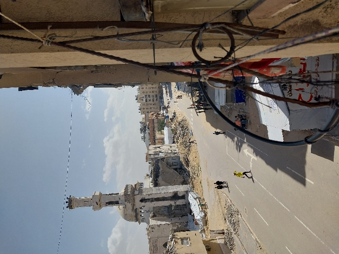 This image depicts a street scene in Khan Yunis, Palestinian Territory, captured from an elevated perspective. The immediate foreground on the right displays a section of a light brown building exterior, from which multiple dark cables and ropes are affixed, some hanging vertically and others running horizontally.
The midground features a paved road with white double lane markings and a single white arrow indicating a turn to the left. A lone individual wearing a bright yellow shirt walks left on the road, casting a pronounced shadow. Further back on the left side of the street, two individuals in dark clothing walk together. On the right side of the street, a group of approximately five people are gathered, some seated, around a white tent-like structure that displays visible Arabic text. Adjacent to this, a smaller, darker vendor stall or tent is present. Additional pedestrians are discernible further along the street in the distance.
The background is characterized by extensive structural damage. Dominating the left side is the Great Mosque of Khan Yunis, identifiable by its white, ornate minaret and dome, both exhibiting severe damage including large breaches and missing sections. Surrounding the mosque and extending through the scene are numerous buildings reduced to rubble, with only skeletal remains and piles of debris visible on dusty, barren ground. Beyond this immediate destruction, a skyline of multi-story buildings, some appearing less damaged, stretches into the distance. The sky above is clear and light blue with sparse white clouds. A thin, dark utility wire runs horizontally across the upper central portion of the frame.