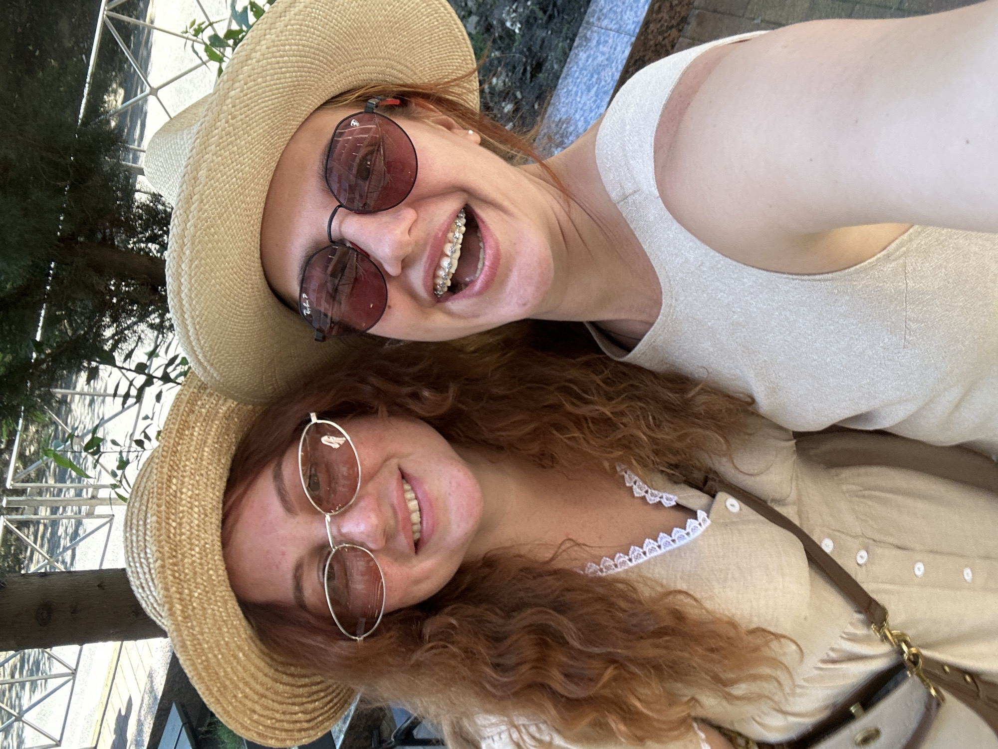 A close-up selfie captured on a bright, sunny day in Sochi, Russia, features two young women smiling broadly. Both are wearing light-colored straw hats and sunglasses.
In the foreground, a woman with long, curly auburn hair and a joyful expression wears round, gold-rimmed sunglasses. "Ray-Ban P" is faintly visible on the right lens. She is dressed in a light beige or cream-colored collared shirt with delicate white lace trim along the neckline, and a brown leather strap from a shoulder bag is visible over her arm. Her smile reveals natural teeth.
Behind her and slightly to the left, another woman with lighter hair and a wide, open-mouthed smile is visible. She wears darker, round-framed sunglasses and reveals dental braces on her upper teeth. Her attire includes a simple, light-colored sleeveless top or tank top.
The background suggests an outdoor setting, possibly a park or garden. Hints of lush green foliage, trees, and a light-colored metal fence or railing are visible, both directly and as reflections in the sunglasses. The bright lighting indicates it is daytime with clear weather. Both individuals appear happy and relaxed, enjoying the sunny environment.
