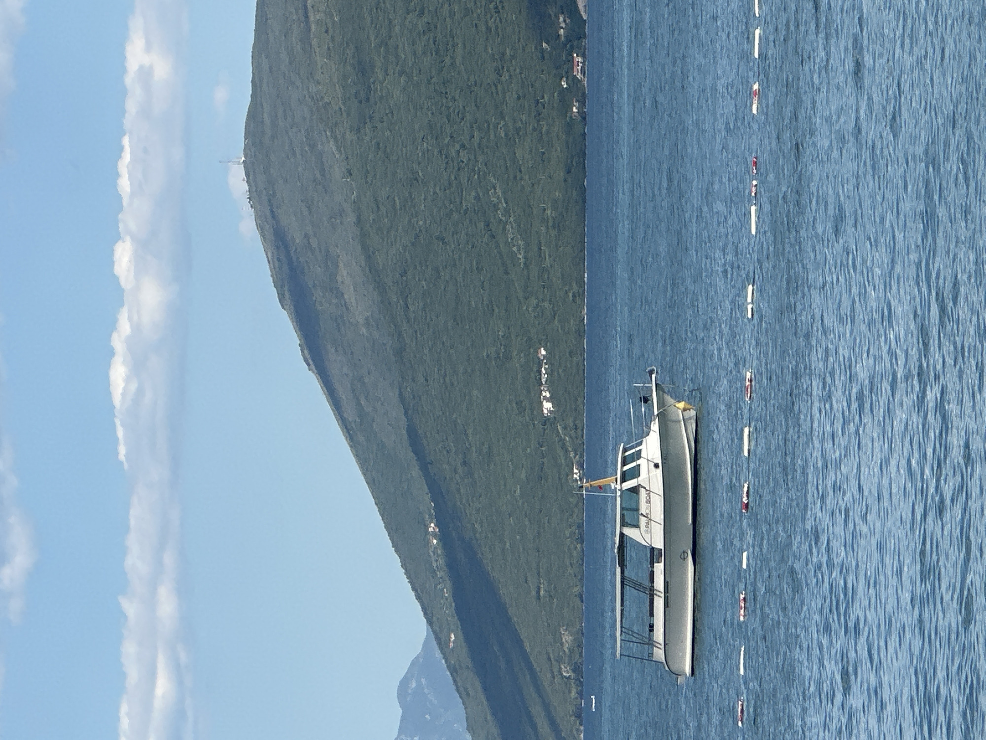 A white motorboat with a canopy is anchored in the blue water of the sea. In the background, a lush green mountain slopes upward, its peak dotted with a tall communication tower and some small buildings. The sky above is a clear blue with scattered white clouds. A row of buoys floats on the water in the foreground, marking a boundary. The scene appears to be the coastline of Igalo, Montenegro.
