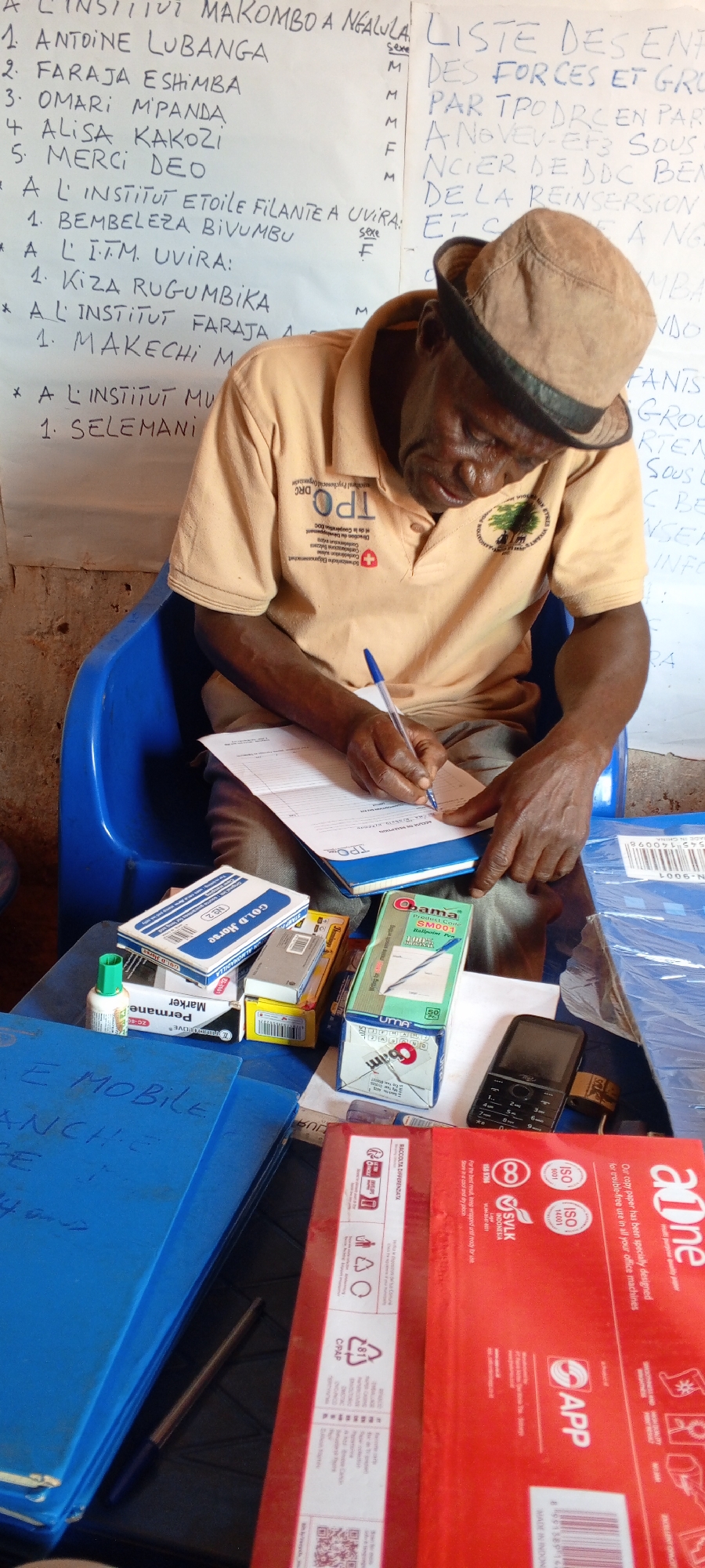 An elderly man wearing a khaki hat and shirt with the logos of "TPODRC" and a tree, sits in a blue plastic chair at a table. He is writing on a form with a blue pen, resting the form on a notebook. On the table are boxes of office supplies, including "Cold Horse" pencils, "Obama" ballpoint pens, and "Permanet Marker," along with a cell phone and a stack of blue folders labeled "E MOBILE ANCHE." A red ream of "aOne" copy paper is also visible. Behind him is a handwritten list titled "A L'INSTITUT MAKOMBO A NGALULA," suggesting the location is the city of Ngalula, Democratic Republic of the Congo. The list appears to be a register of names and genders ("Sexe") related to "LISTE DES ENF DES FORCES ET GRO PAR TPODRC EN PART A NOVEV-EF3 SOUS NCIER DE BOC BEN DE LA REINSERSION ETC." The scene suggests an administrative or record-keeping activity related to child soldiers or reintegration efforts.