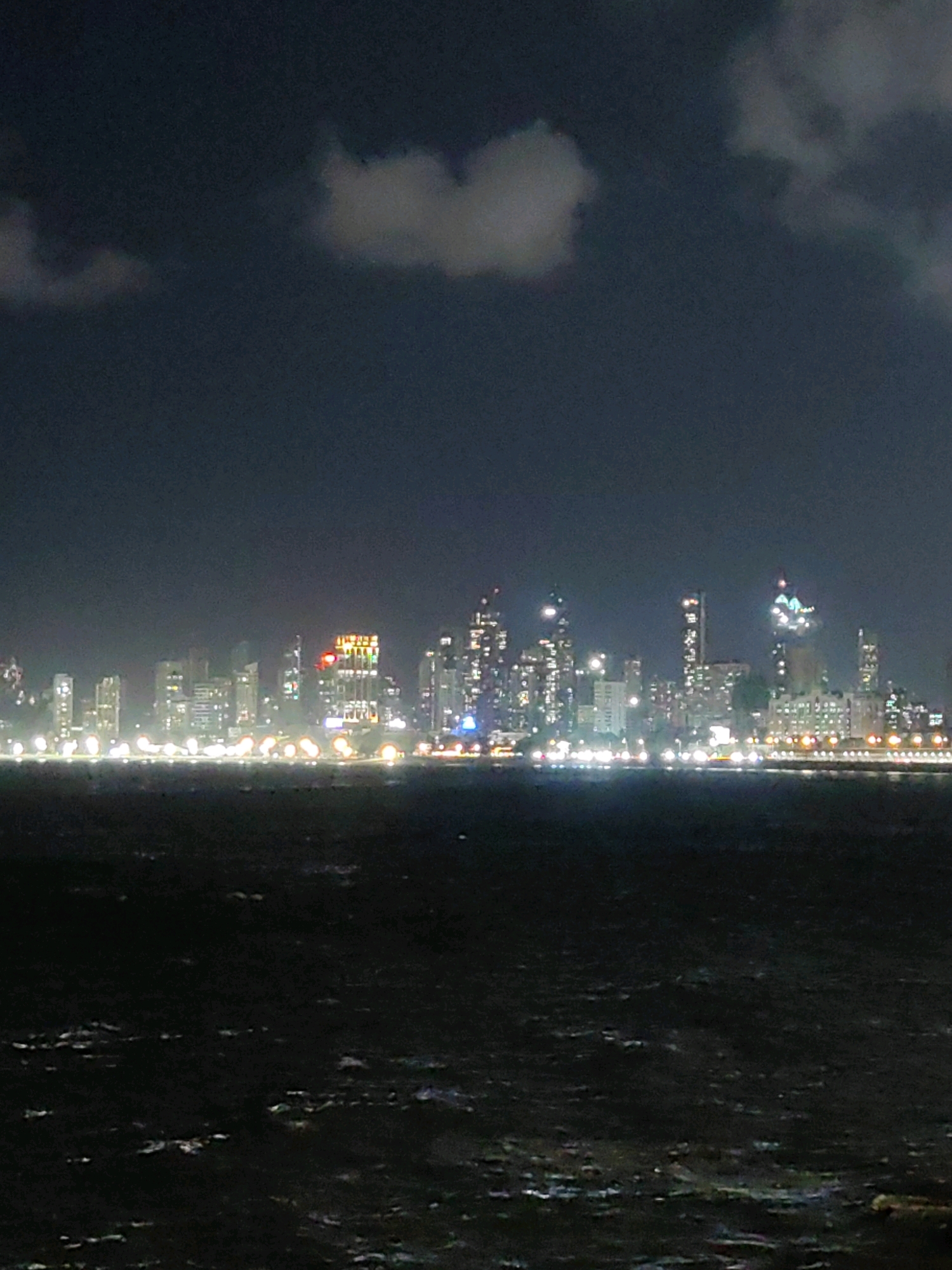 An image captures a nighttime view of the Mumbai skyline, likely from across a body of water. The dark water in the foreground has ripples and reflects some of the city lights. The cityscape is dominated by numerous tall buildings, many illuminated with white lights. Some structures feature distinctive colored lights, including a building with prominent yellow and red lights, suggesting it may be a hotel or commercial establishment. The sky above is dark, with a few faint clouds visible, also catching some ambient light from the city. The overall impression is a vibrant urban landscape at night, viewed from a distance.
