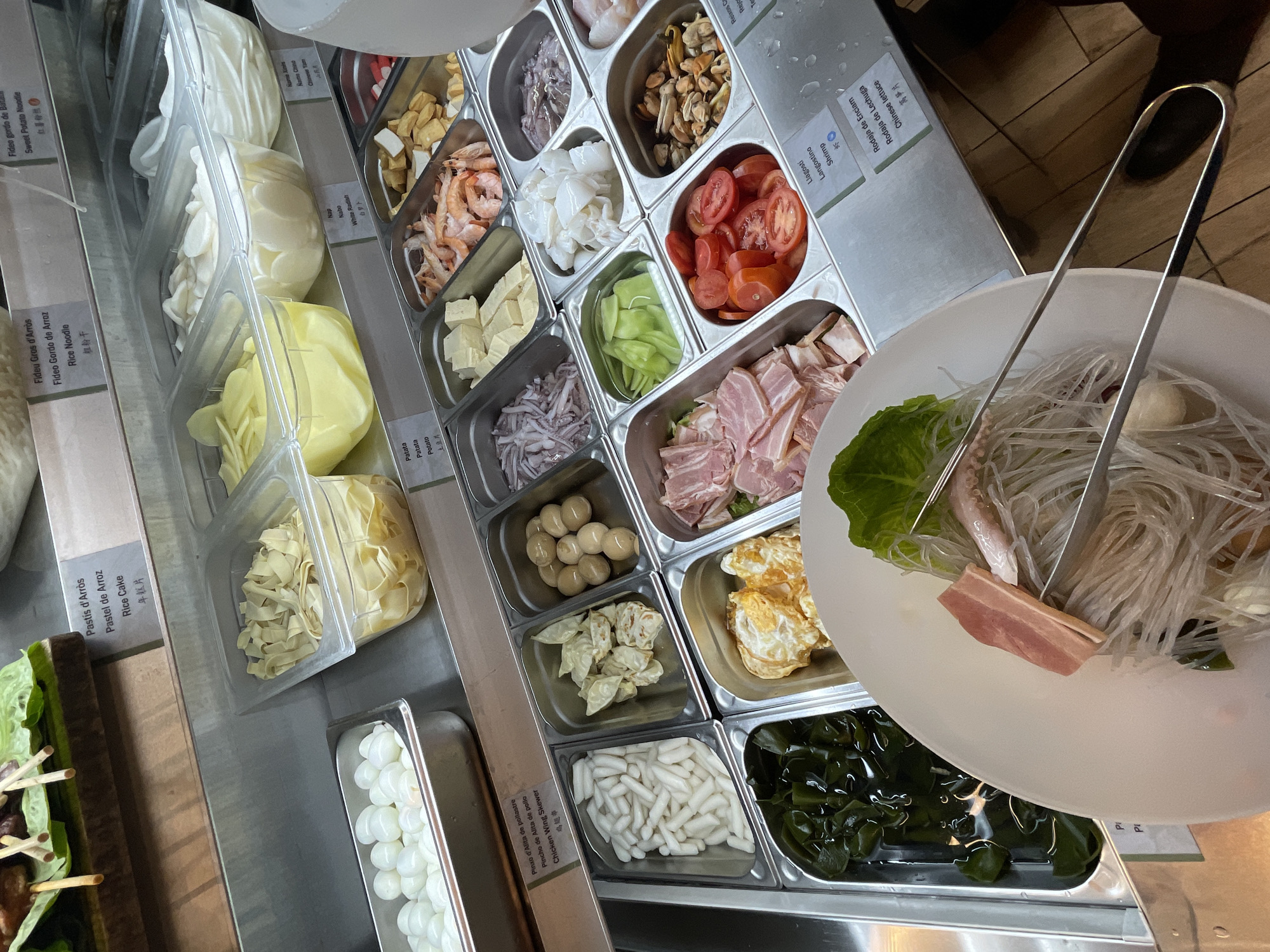 This is a photograph taken at a self-service food station. The items are contained within stainless steel trays. The trays hold different ingredients that can be added to a food dish. The ingredients appear to be a variety of noodles, vegetables, seafood, meats, and possibly eggs. A white bowl contains some of the ingredients, including lettuce, noodles, and a piece of bacon. A pair of stainless steel tongs are also present, seemingly used for selecting and adding items to the bowl. There is no indication that this is in Barcelona, Spain.