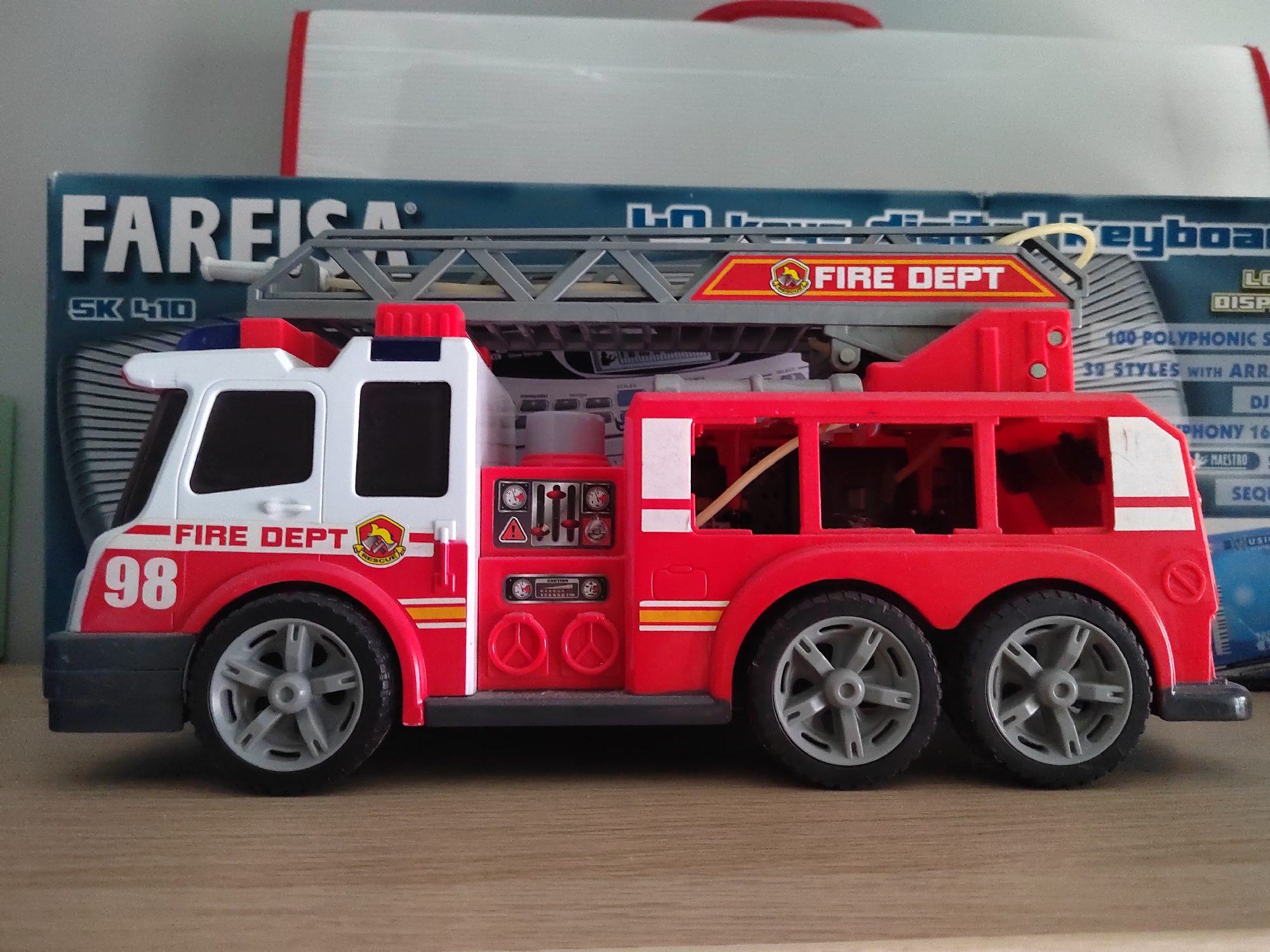 A toy fire truck with the number 98 on its side is parked on a wooden surface. The fire truck is red and white and has a ladder on top. The truck is facing to the left. In the background, there is a box that says "FAREISA" and "KEYBOARD". The scene is indoors and appears to be in a residential setting, possibly in Porto Recanati, Italy, given the presence of the "FAREISA" branded box, which is a common electronics retailer in Italy.