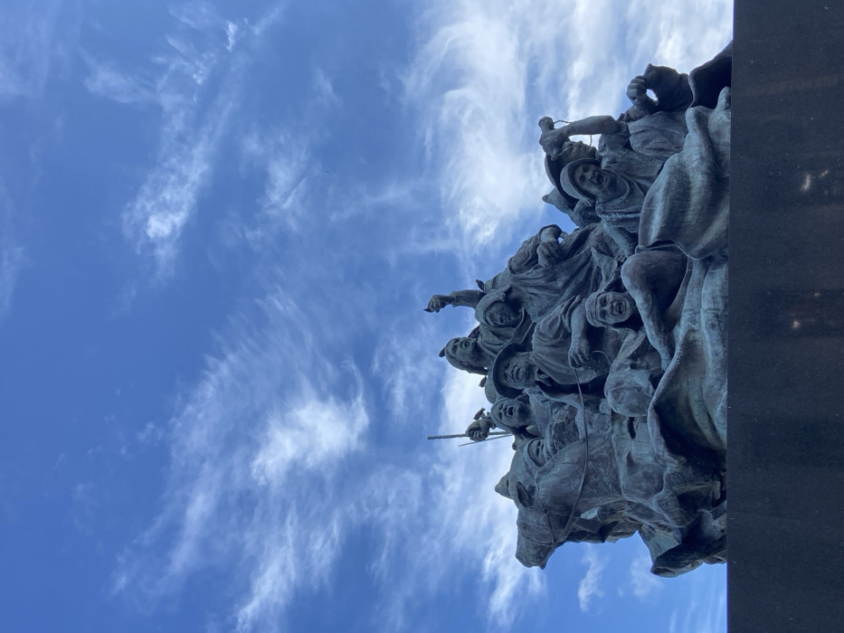 A bronze sculpture depicts a chaotic scene of figures, some on horseback, against a bright blue sky with wispy white clouds. The figures are expressive, with strained faces and dynamic poses. The sculpture appears to be part of a monument, with a dark, vertical structure visible on the right. The low-angle shot emphasizes the sculpture's height and dramatic composition.