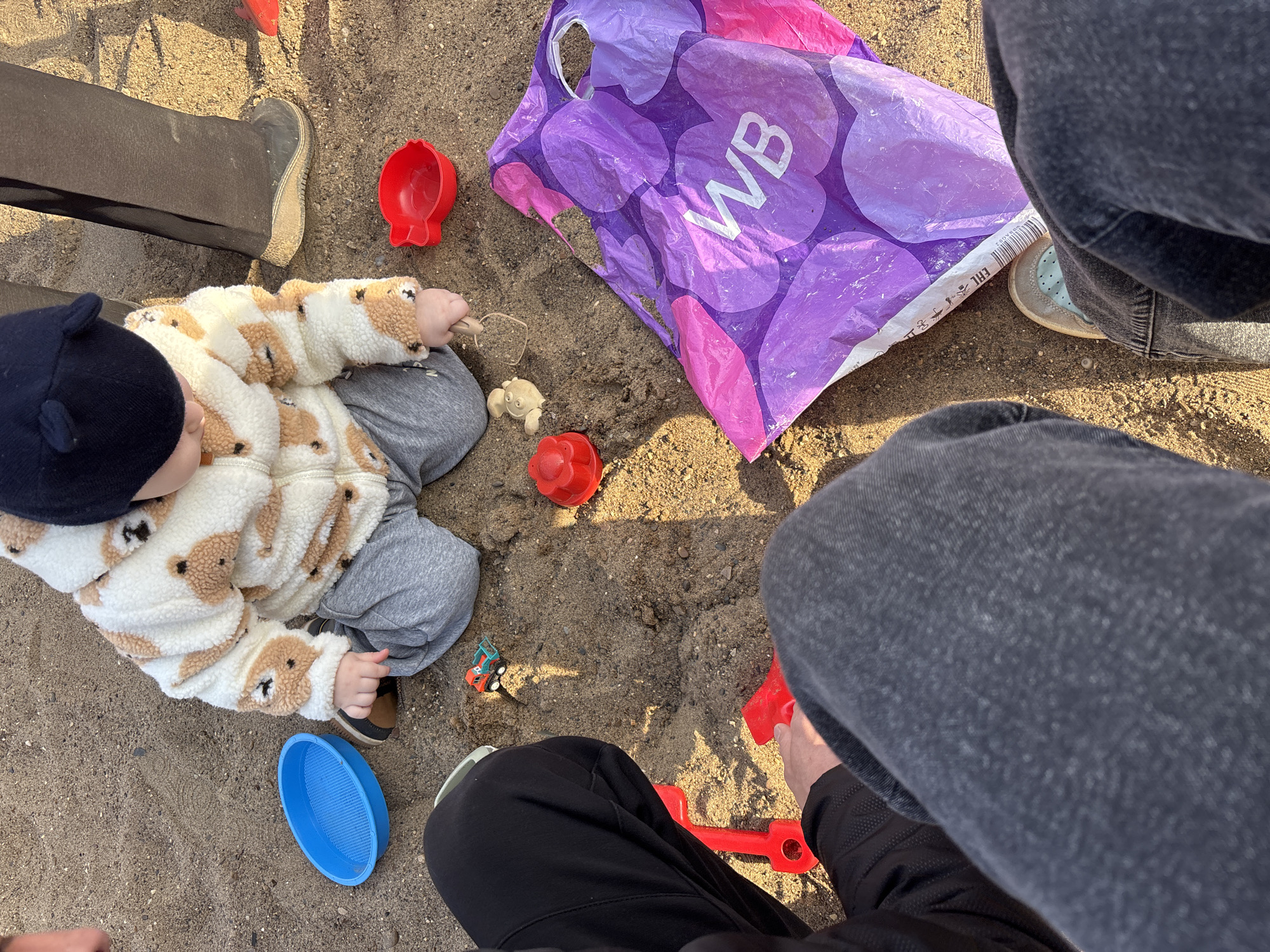The image shows an outdoor scene, presumably a playground, with sand covering the ground. A toddler in a teddy-bear-patterned jacket and a dark blue hat sits on the sand, playing with various colorful plastic toys, including a red cup, a blue sieve, and a small truck. Beside the toddler is a bag with a WB logo on it. Two adults are partially visible, with their legs and lower bodies in the frame, indicating they are sitting or kneeling nearby, likely accompanying the child. One adult is wearing grey jeans and the other black trousers and a jacket. There is no identifiable information related to the city of Chaikovsky, Russia.