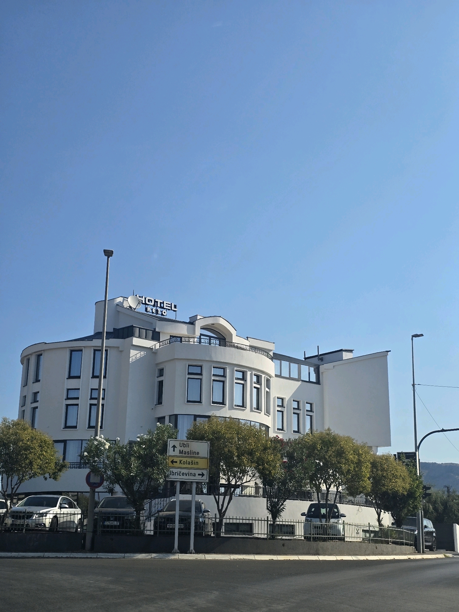 The image shows a modern white hotel named "HOTEL KETO" in Podgorica, Montenegro, under a clear blue sky. The hotel features a curved facade on the left with multiple windows and a section with a balcony. In front of the hotel, there are several cars parked behind a fence, and trees with green foliage line the property. A street signpost is visible with directions to "UBLI Masline", "Kolašin", and "Ibricovina". The foreground shows a dark asphalt road.