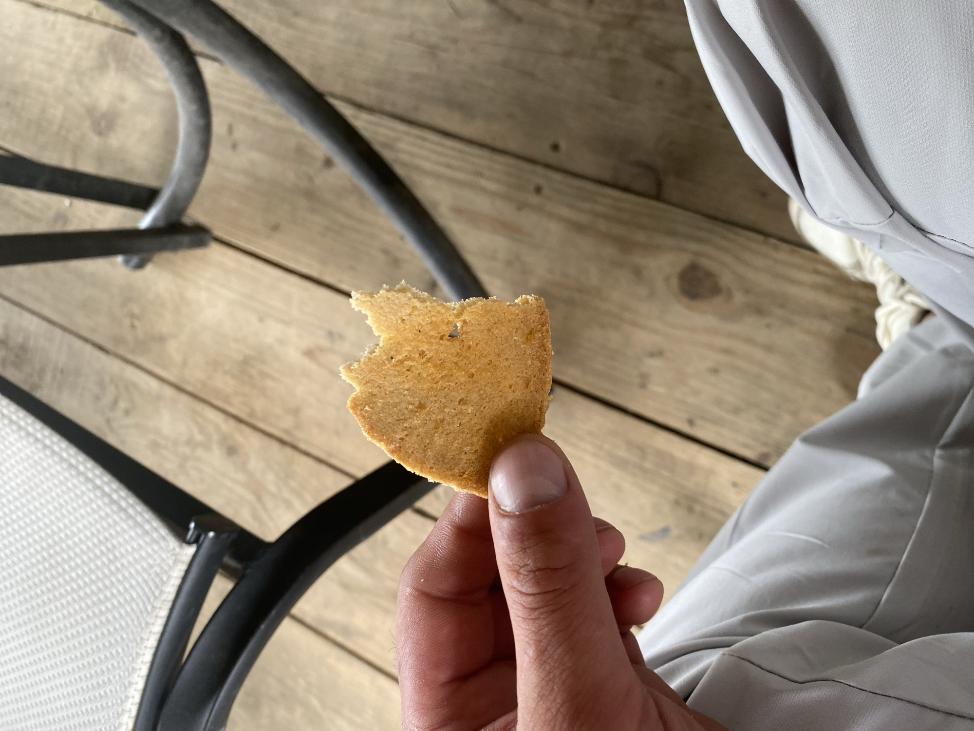 A close-up shot captures a person's hand holding a broken piece of a cookie over a wooden surface. The cookie is light brown and appears to be slightly crumbly. The hand, with its fingernails visible, is positioned in the lower right portion of the frame.
In the background, wooden planks form a textured floor, with visible grain and subtle variations in color. To the left, a portion of a black metal chair is visible, with its curved legs and a part of the seating area, which is covered in a light-colored, possibly fabric, material. Further to the right, the person is wearing light-colored pants, likely made of a smooth fabric, with soft folds and creases. A white shoe is also partially visible, suggesting the person is seated or leaning.
The lighting in the image suggests it is daytime, with natural light illuminating the scene. The overall atmosphere is casual and relaxed, possibly indicating a moment of taking a break or enjoying a snack outdoors. No text is visible in the image. The location context of Balta, Ukraine, does not provide specific visual cues within the image itself, but suggests an outdoor setting, potentially a patio or balcony.