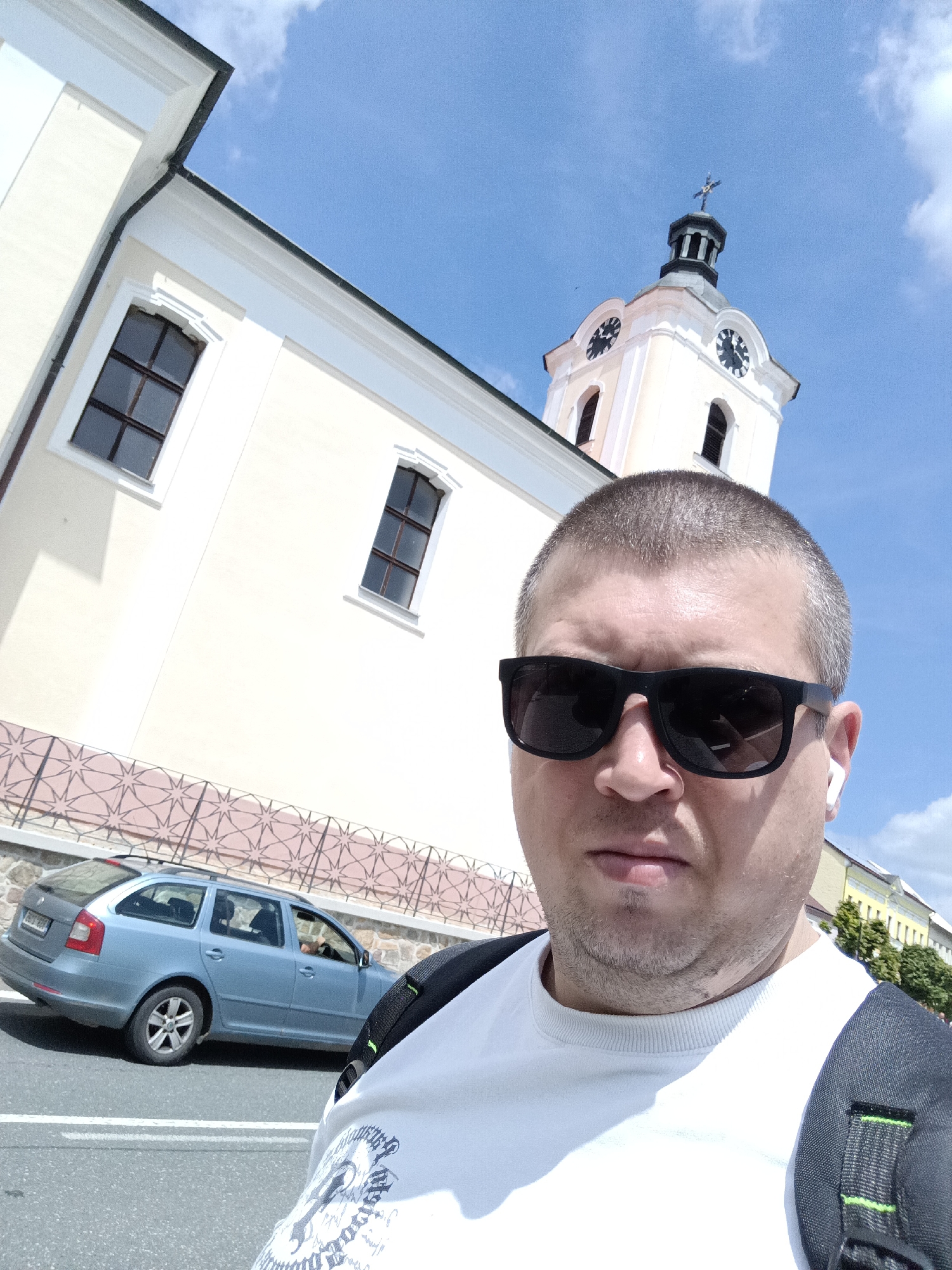 A man is taking a selfie outdoors in front of a light yellow building with a clock tower. He is wearing sunglasses and a white t-shirt with a backpack. A blue station wagon is parked on the street to his left. The building appears to be a church or similar public structure with arched windows. The sky is clear with a few white clouds. The scene is consistent with a location in a European town, possibly in Dalovy, Czechia.