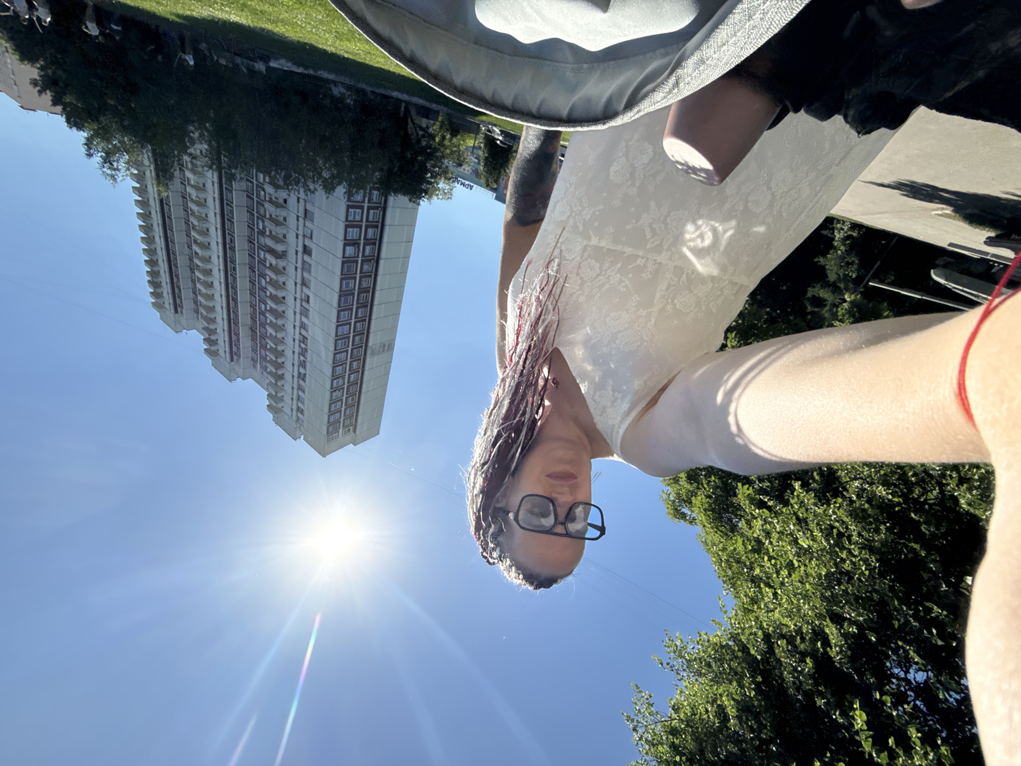 A low-angle outdoor photograph captures a woman in a white dress taking a selfie or wide-angle shot from a stroller's perspective on a sunny day in Moscow, Russia. The woman, positioned centrally, has fair skin, dark hair with red/pink highlights styled in braids or dreadlocks, and wears dark-framed sunglasses. A red string bracelet is visible on her left wrist, and tattoos cover her right arm. Her expression shows a slight pout. In the immediate foreground, the light-gray interior edge of a stroller or baby carriage is visible, containing a pink bottle or cup. To the left, a dense canopy of green trees borders a paved path, behind which a dark, large object resembling a tank or military vehicle is partially discernible. The background is dominated by a clear, bright blue sky with a prominent sun causing lens flares. Against the sky on the right are tall, multi-story residential buildings, characteristic of Soviet-era architecture, featuring numerous windows and balconies. A Cyrillic sign displaying "АРМАЯ" is faintly visible in the mid-background. The presence of military vehicle exhibits alongside residential buildings and a park-like setting suggests the location is in the vicinity of Victory Park (Парк Победы) in Moscow.