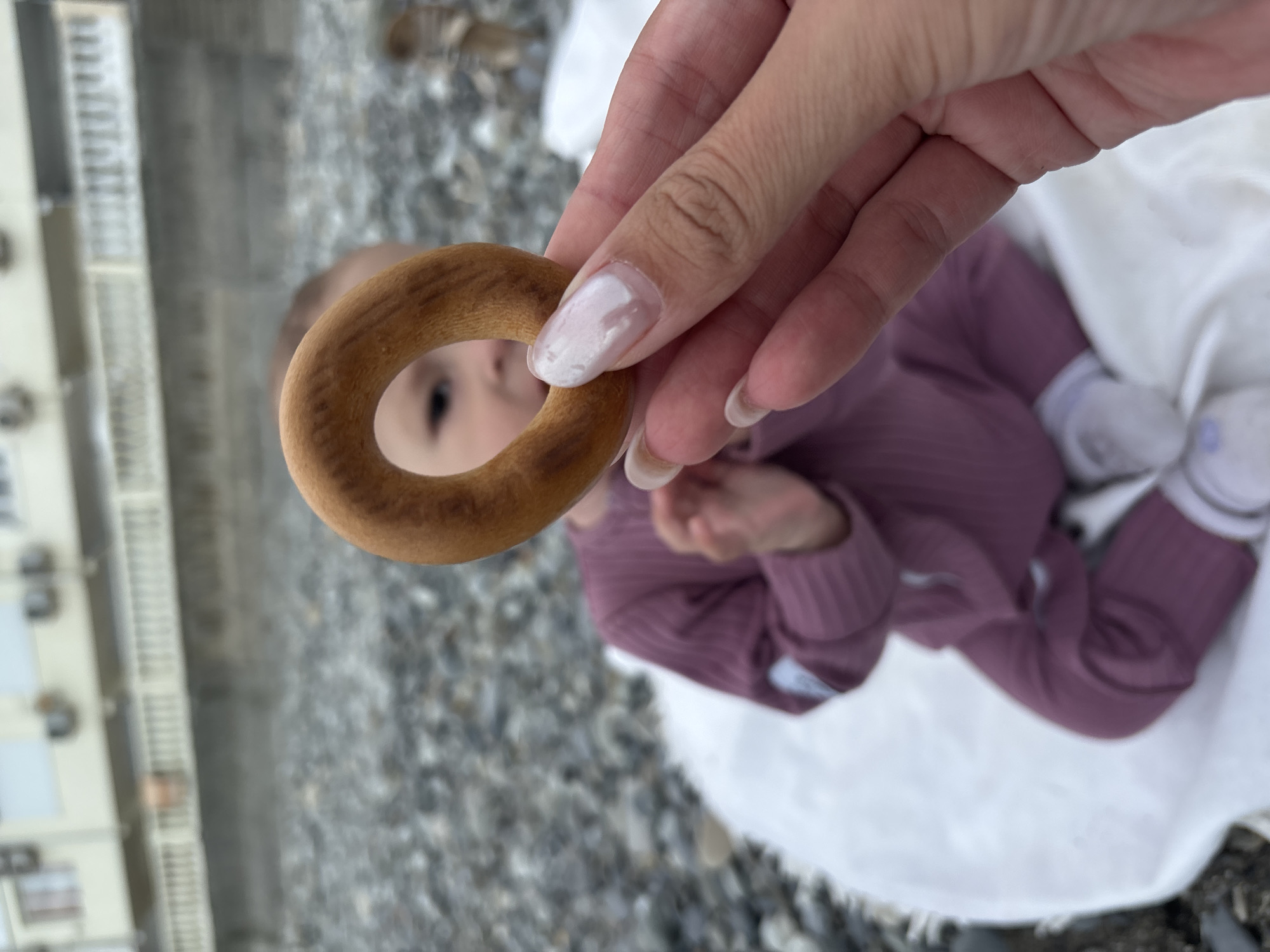 A human hand holds up a ring-shaped bread product against the backdrop of a pebble beach, with a blurred infant visible through the hole in the bread. The infant, wearing a purple outfit and white socks, lies on a white cloth. In the background, a building can be seen on the left side of the frame. The picture was taken in Sochi, Russia.
