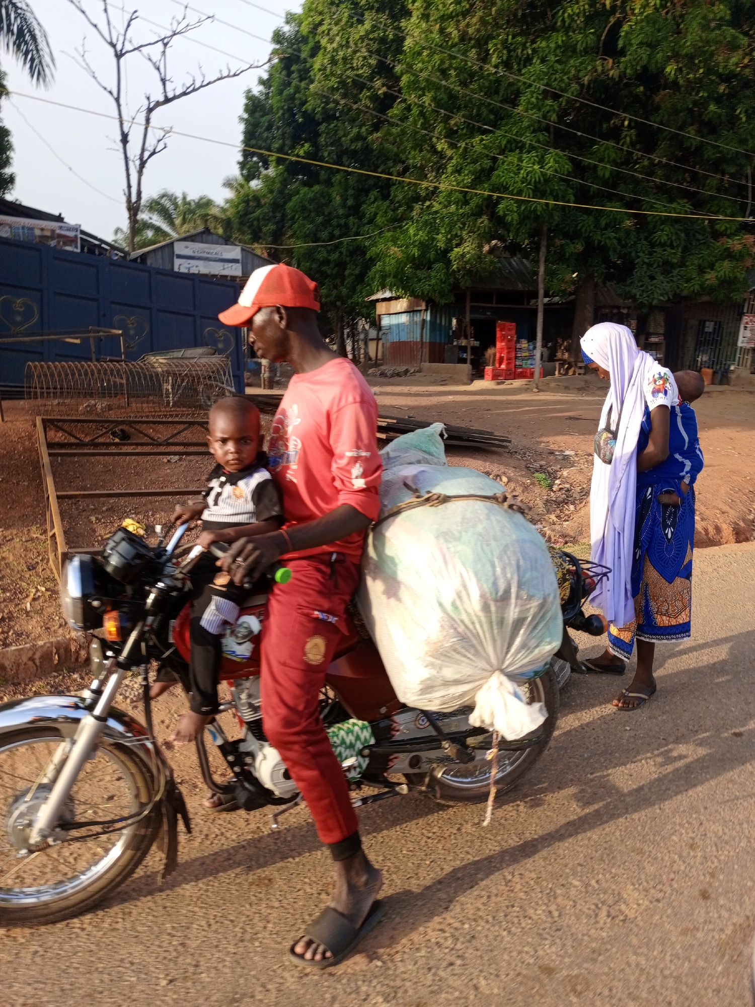 The image depicts a man on a red motorcycle in Keffi, Nigeria. He is wearing a red shirt, red pants, a red and white baseball cap, and black sandals. A small child is seated in front of him on the motorcycle. A large, white bag is strapped to the back of the motorcycle. A woman wearing a long white head covering and a blue dress with a child secured on her back is standing nearby. In the background are trees, buildings, and a blue metal fence. The road is paved and the scene appears to be during daylight.