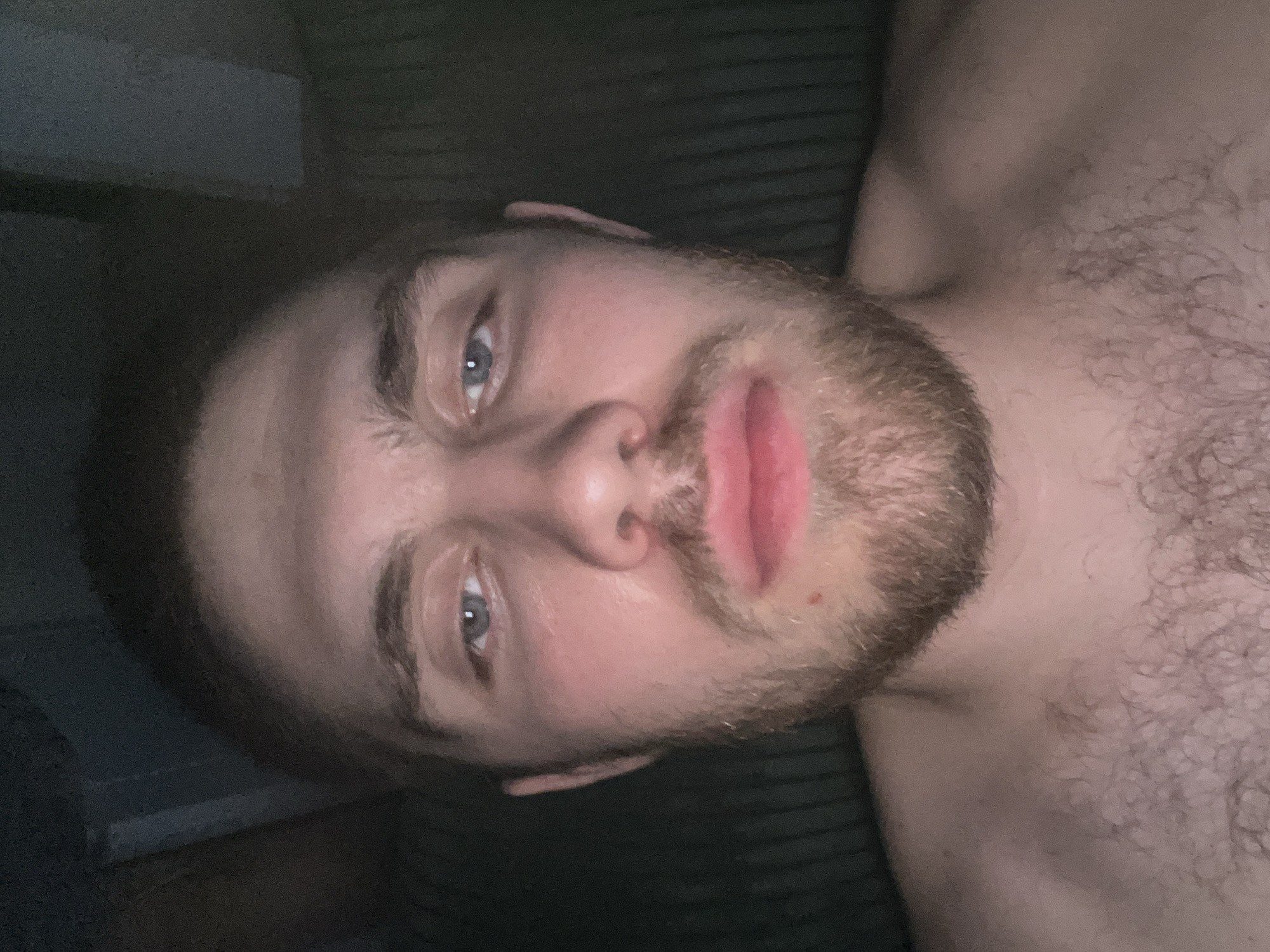The image is a close-up, head-and-shoulders selfie of a light-skinned male with blue eyes, brown hair, and a beard. He is shirtless and appears to be indoors, with a dark, textured background that could be a wall or furniture. The lighting is dim, casting shadows on his face and highlighting his features. There is a slight redness on his chin and cheeks, possibly due to lighting or skin tone. The man's expression is neutral. The image does not contain any information that would allow for the identification of a specific location within Pruszcz Gdański, Poland.