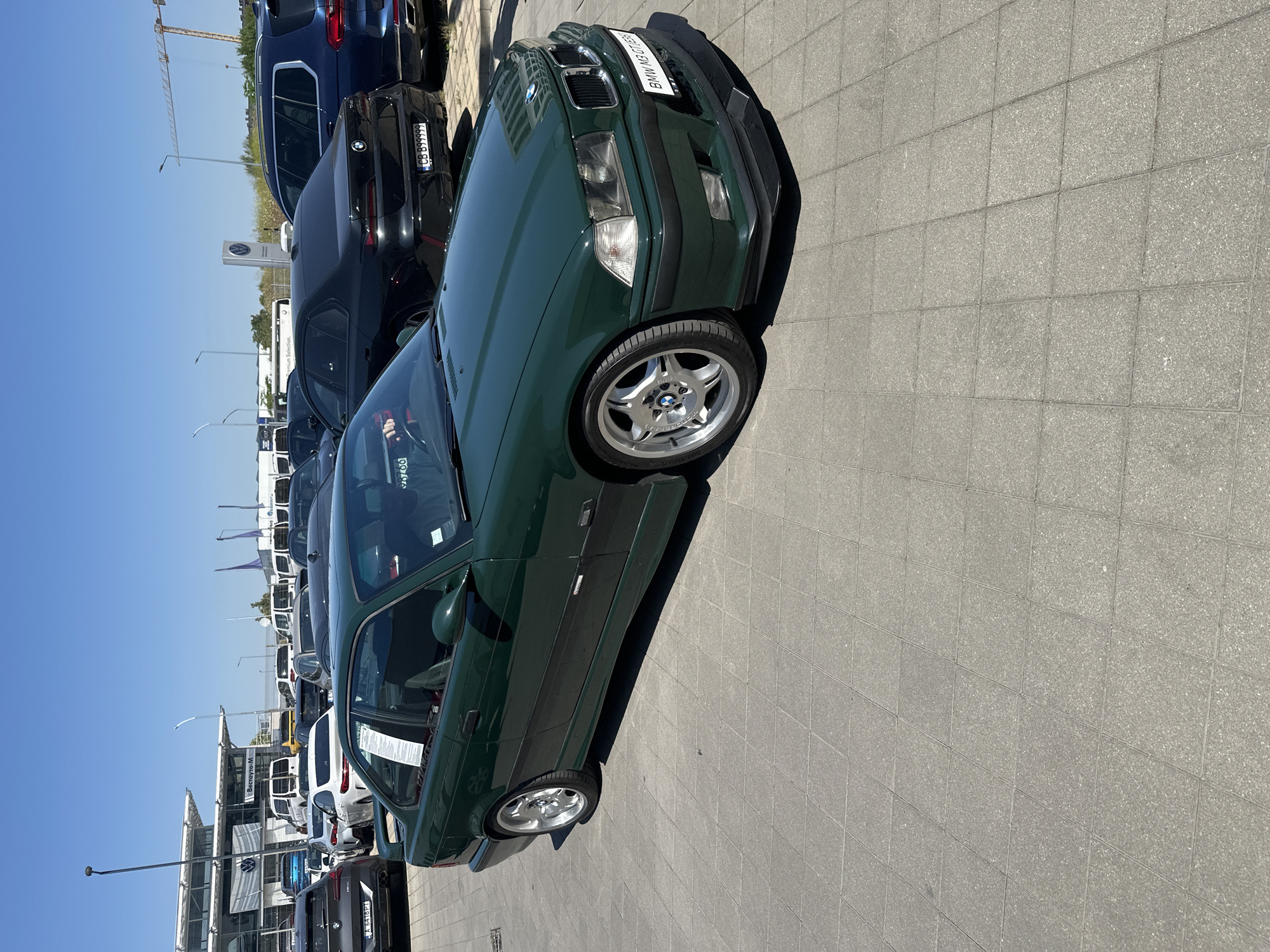 A dark green BMW M3 coupe is parked outdoors on a sunny day, with its front passenger side closest to the camera. The car is angled, with its front wheels turned slightly to the left. The car has a black front bumper and black side trim. The wheels are silver with a five-spoke design. A white license plate is visible on the front of the car, with text that appears to read "BMW M3 GT".
In the background, several other cars are parked in a lot. The cars are of various colors and models, including a dark blue SUV and a black sedan with a license plate that reads "GB B99999". Further in the background, there are buildings and a construction crane. The ground is paved with square gray tiles. The sky is clear and blue, indicating a sunny day. The overall scene suggests a car dealership or a parking lot where cars are on display. No people are visible in the image.